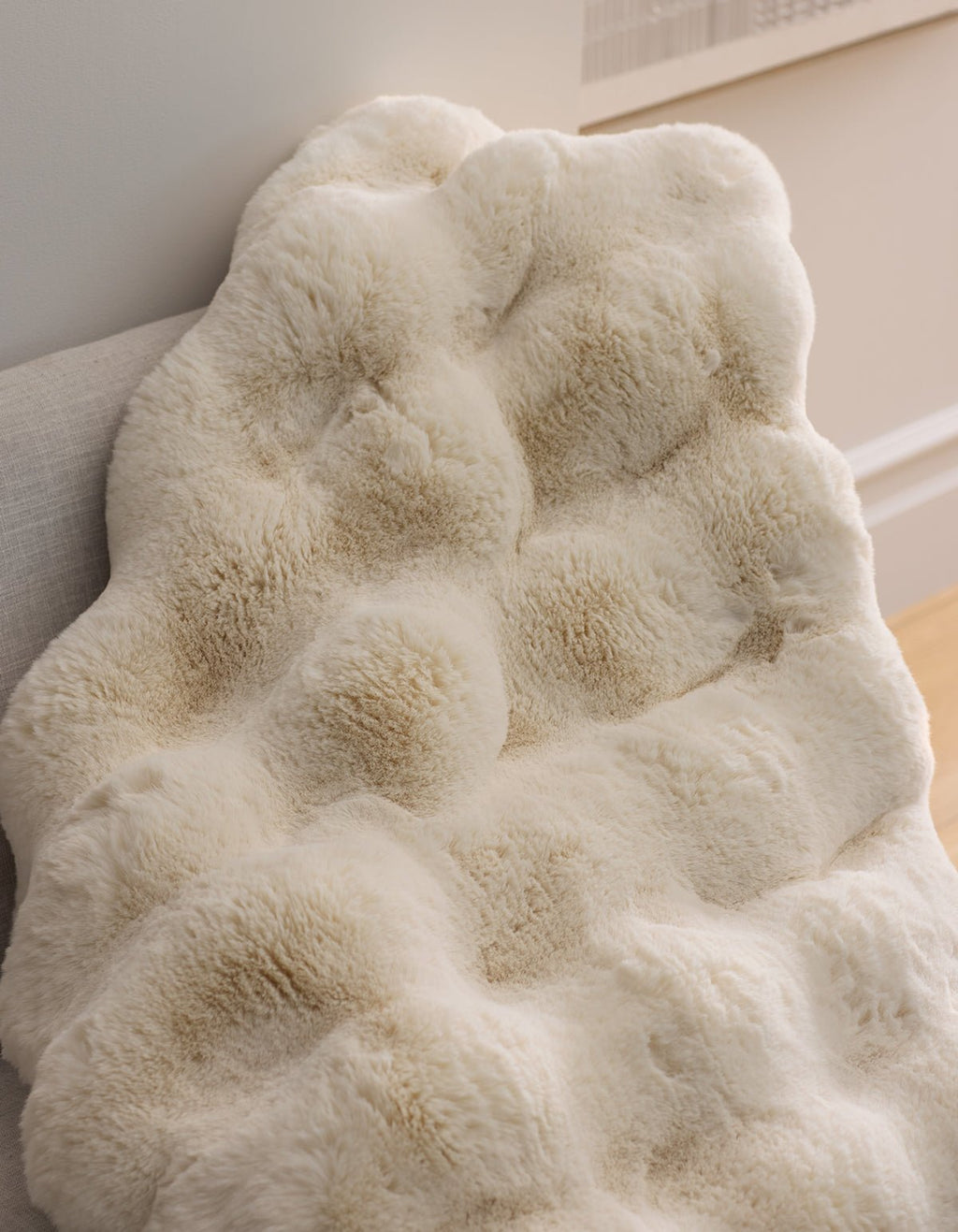 The Cozy Earth Double Bubble Cuddle Blanket, featuring a cream-colored faux fur with a textured, tufted design, is draped over a light chair in a stylish, comfortable room.|Color:Creme