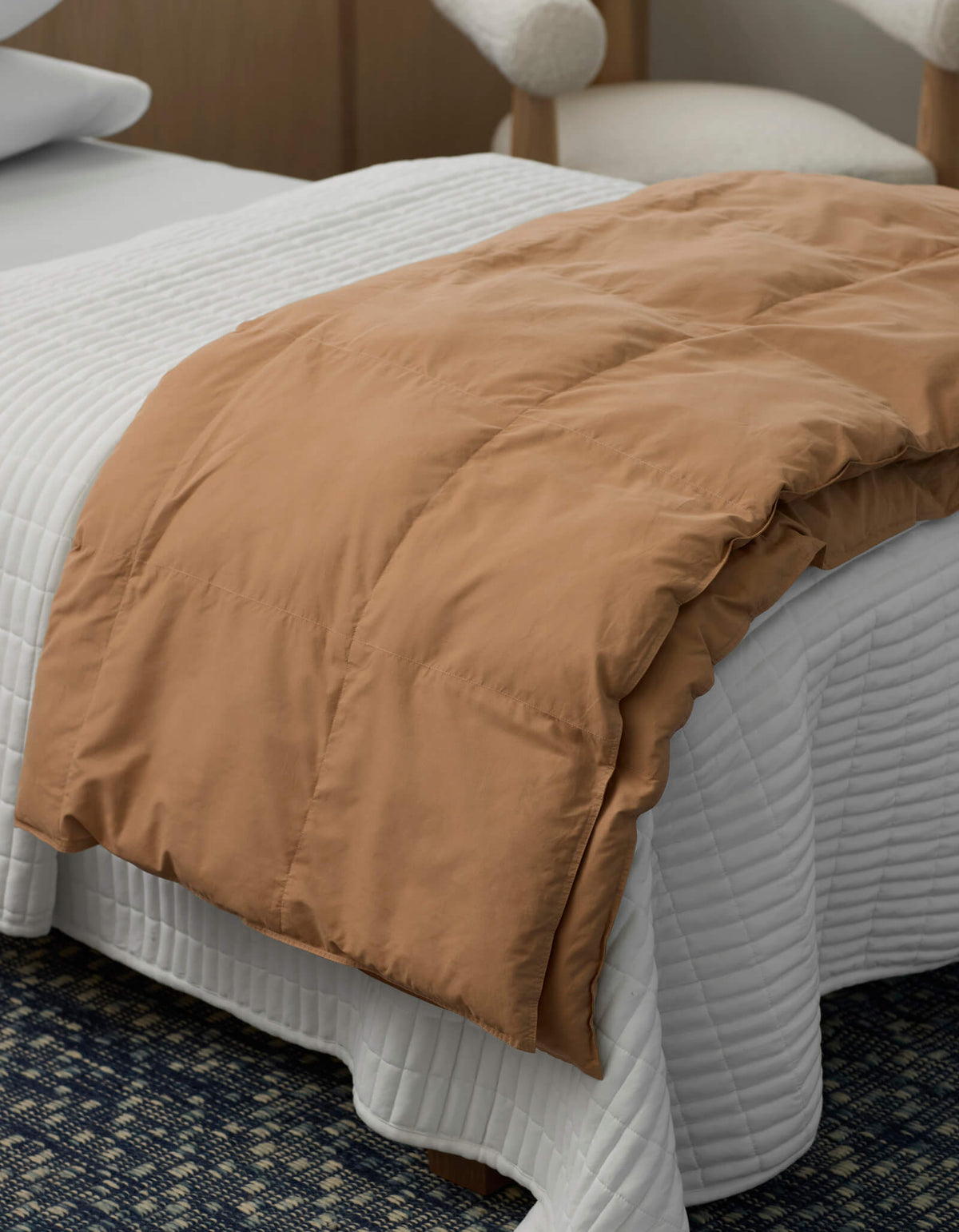 A neatly made bed features a Cozy Earth Down Alternative Throw in white, quilted and draped across the foot. In the background, a modern wooden chair with a white cushion is visible.|Color:Sandalwood