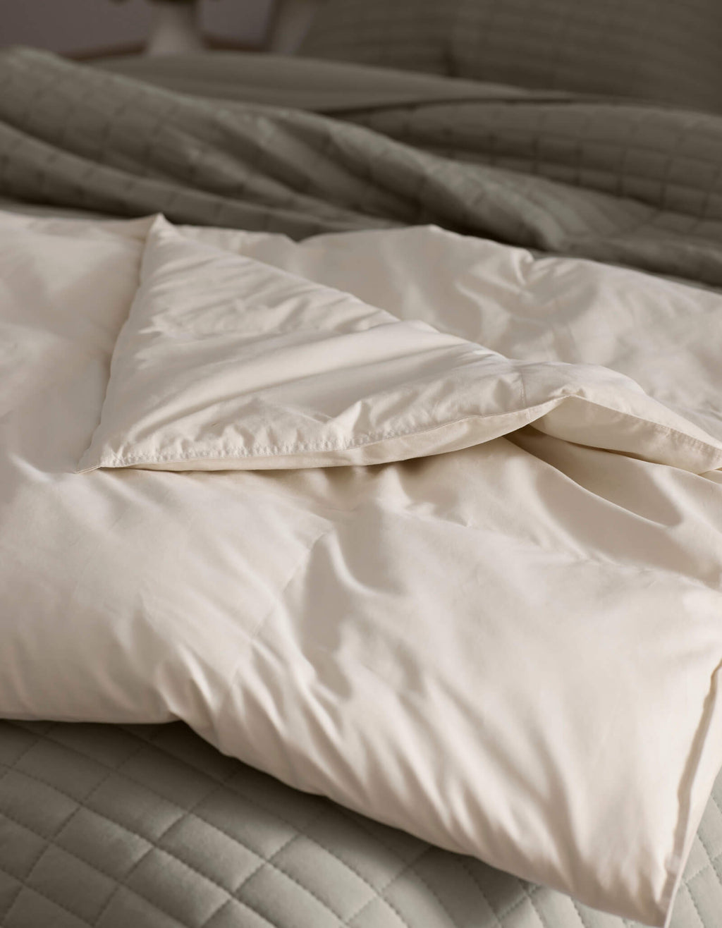 A close-up of Cozy Earth's Down Alternative Throw, folded atop a quilted light gray bedspread. The soft, cream-colored fabric looks cozy and inviting, with natural light highlighting its smooth texture.|Color:Coconut