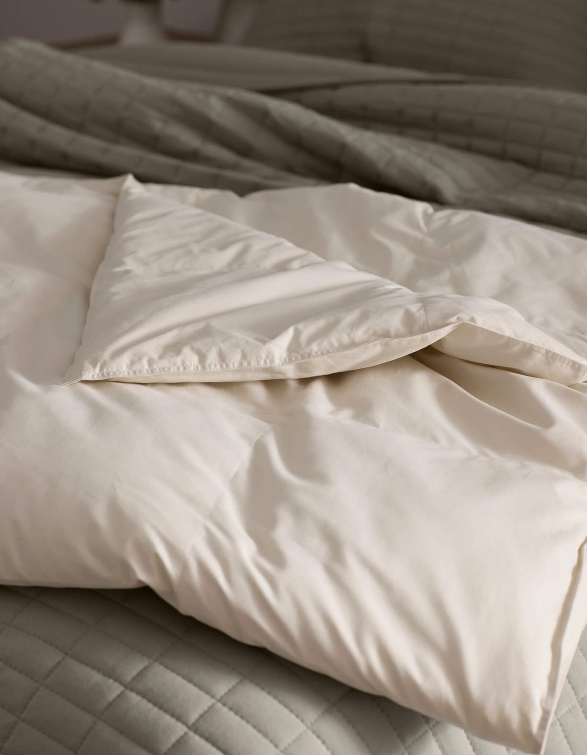 A close-up of Cozy Earth's Down Alternative Throw, folded atop a quilted light gray bedspread. The soft, cream-colored fabric looks cozy and inviting, with natural light highlighting its smooth texture.|Color:Coconut