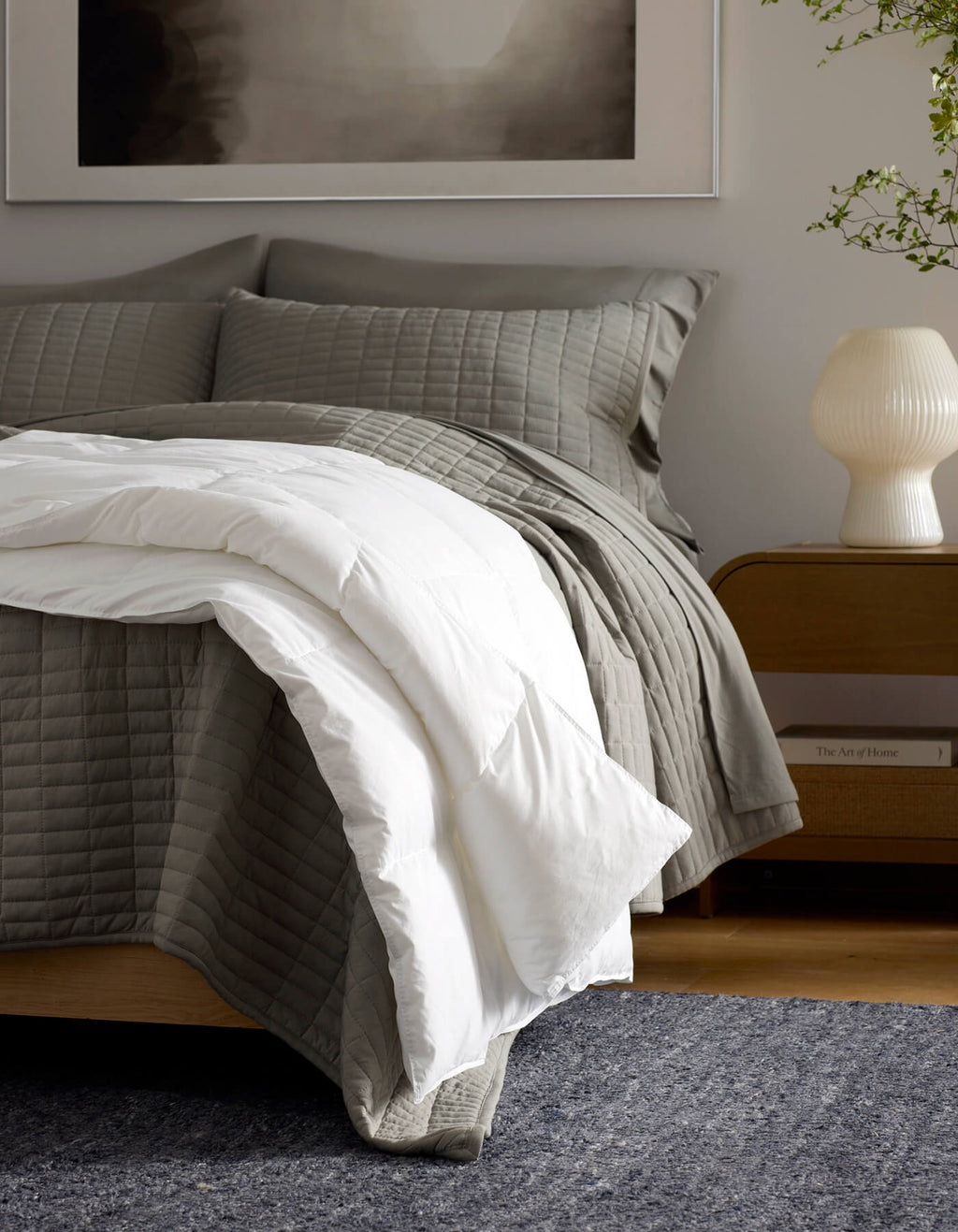 A cozy bedroom features a neatly made bed with gray quilted bedding and the Cozy Earth Down Alternative Throw, a wooden nightstand with a white lamp, and a plant in the background.|Color:White