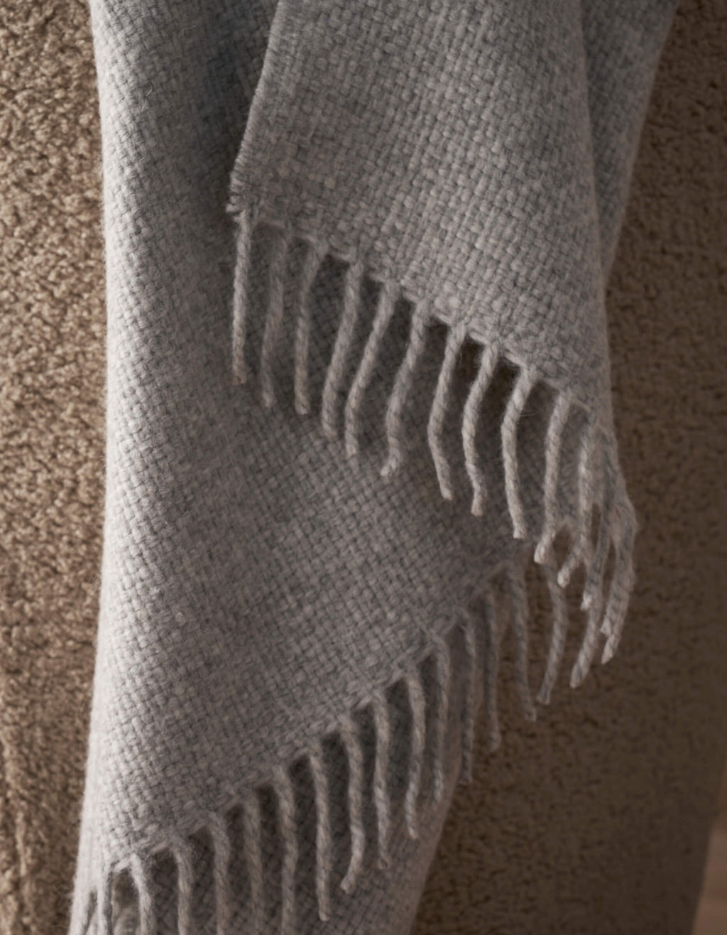 A close-up of the Lucia Throw by Cozy Earth, a soft, textured gray blanket with fringe edges, draped over a coarse tan upholstered surface.|Color:Soft Grey