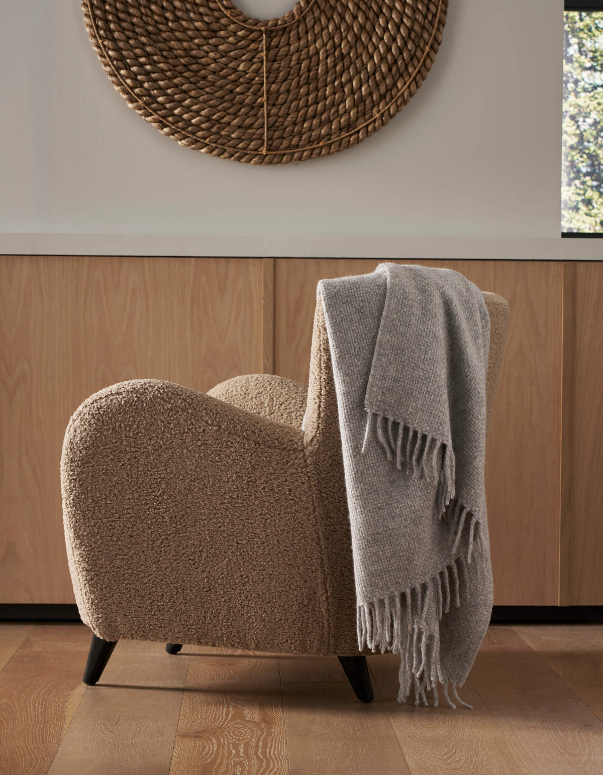 A cozy beige armchair with the Cozy Earth Lucia Throw in soft gray draped over its side sits on a wood floor. Behind it are light wooden cabinets and a round woven wall decor, with sunlight streaming through a nearby window.|Color:Soft Grey