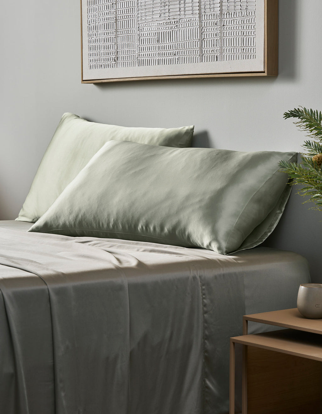 A neatly made bed features Cozy Earth Silk Pillowcases in light sage green, complemented by a wooden nightstand with a plant and cup. A framed abstract artwork decorates the light gray wall above.|Color:Laurel