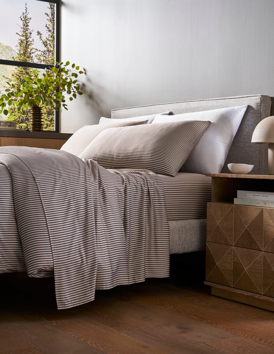 A bed with a Cozy Earth Bamboo Duvet Cover in gray and white stripes sits by a wooden nightstand topped with books and a lamp. Natural light streams in from a window where a potted plant rests on the sill, revealing trees outside.|Color:Classic Stripe in Ebony