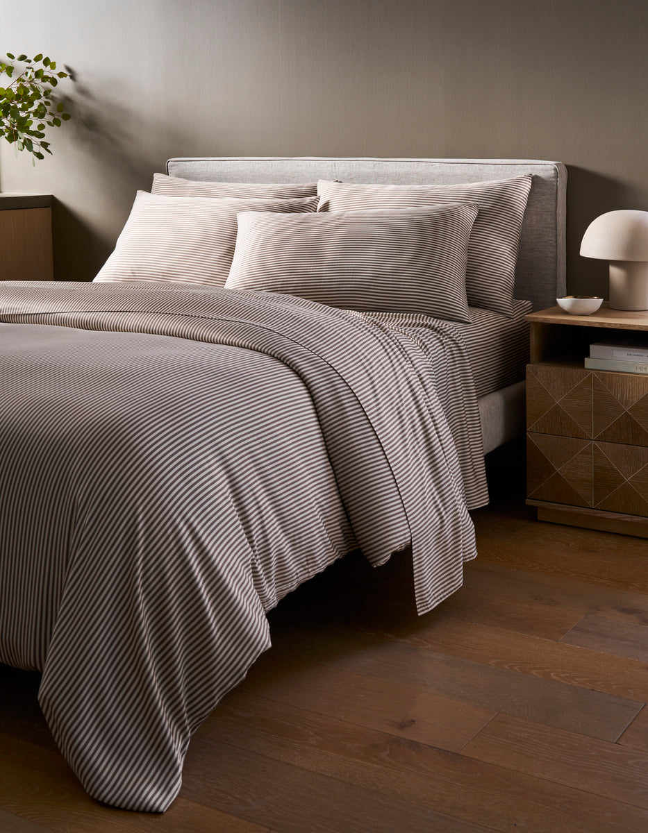 A neatly made bed with the Cozy Earth Bamboo Duvet Cover in gray and white stripes stands in a modern bedroom with wooden floors, a nightstand, a lamp, and a small plant in the corner.|Color:Classic Stripe in Ebony
