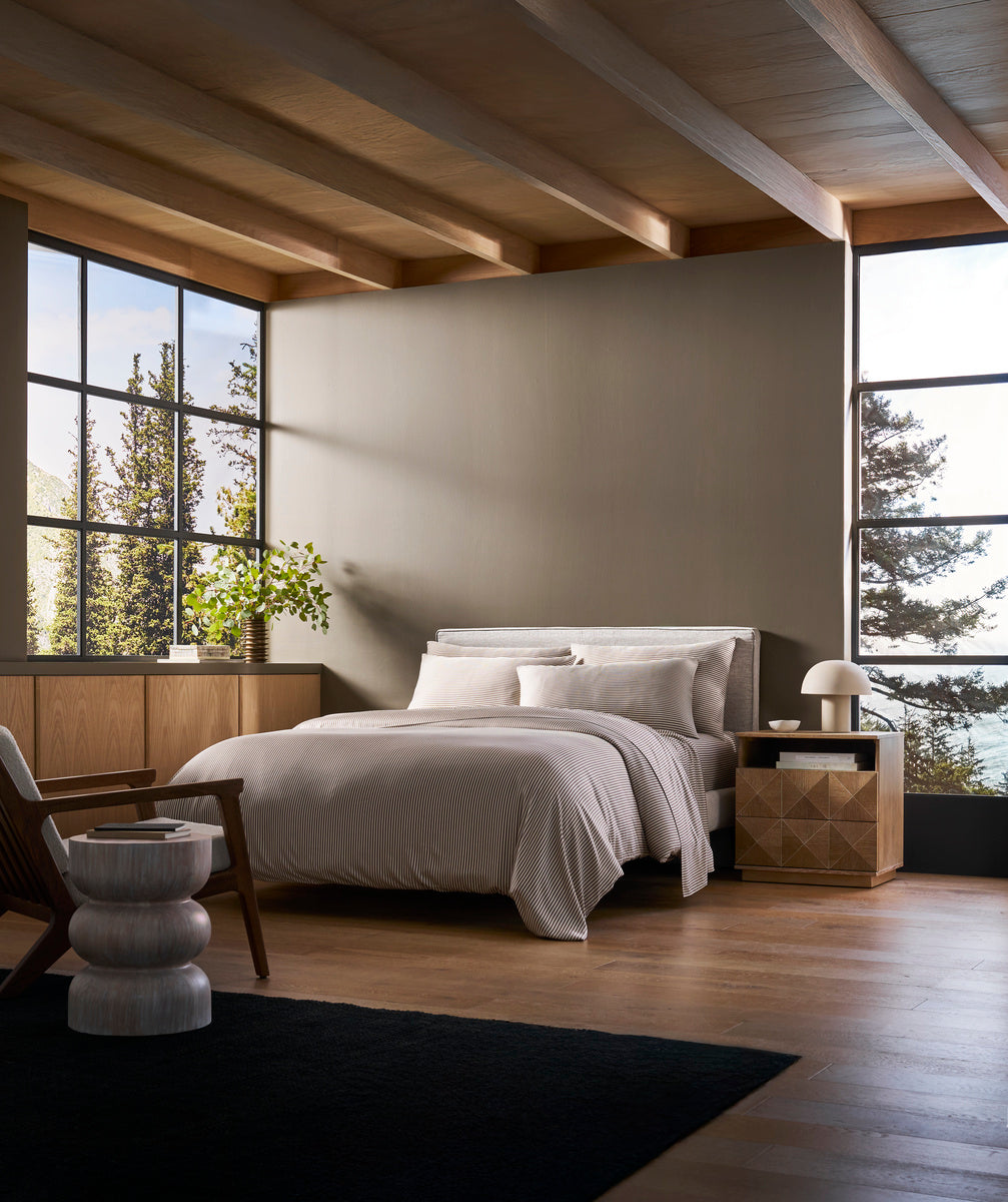 A modern bedroom with large windows, wood floors, a bed featuring the Cozy Earth Bamboo Duvet Cover in white, a wooden nightstand, lamp, black rug, a unique round stool, and views of trees outside.|Color:Classic Stripe in Ebony