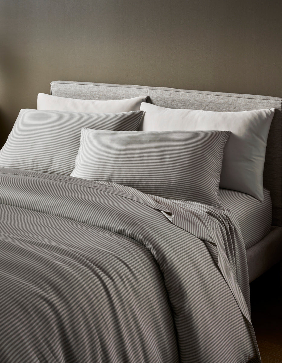 A neatly made bed with a light gray upholstered headboard, white pillows, and the Cozy Earth Bamboo Duvet Cover in gray stripes sits in a softly lit room with neutral walls.|Color:Classic Stripe in Laurel