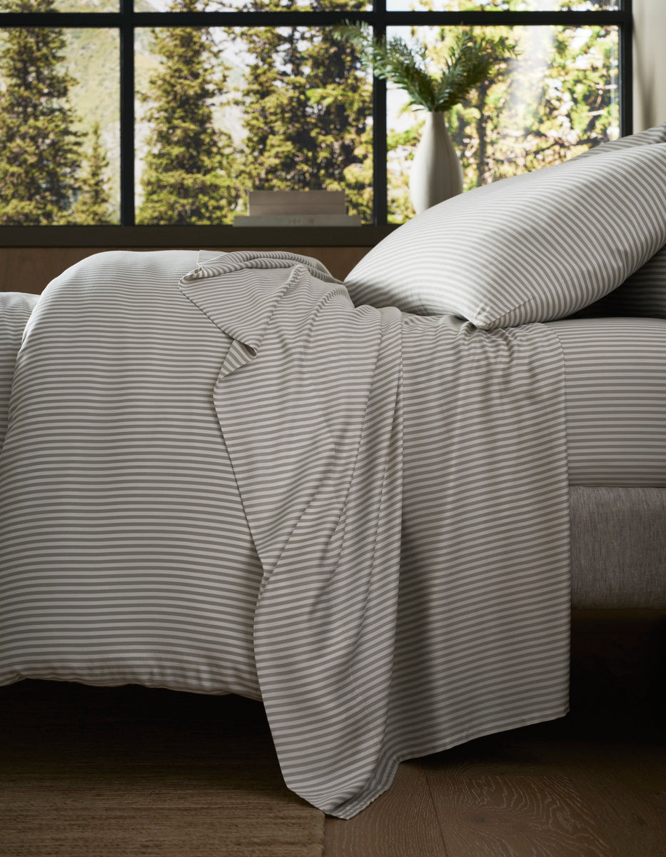 A neatly made bed with the Cozy Earth Bamboo Bedding Core Bundle in gray and white stripes sits by a large window, sunlight streaming in to illuminate a vase of green foliage on the windowsill and reveal views of trees outside.|Color:Classic Stripe in Laurel