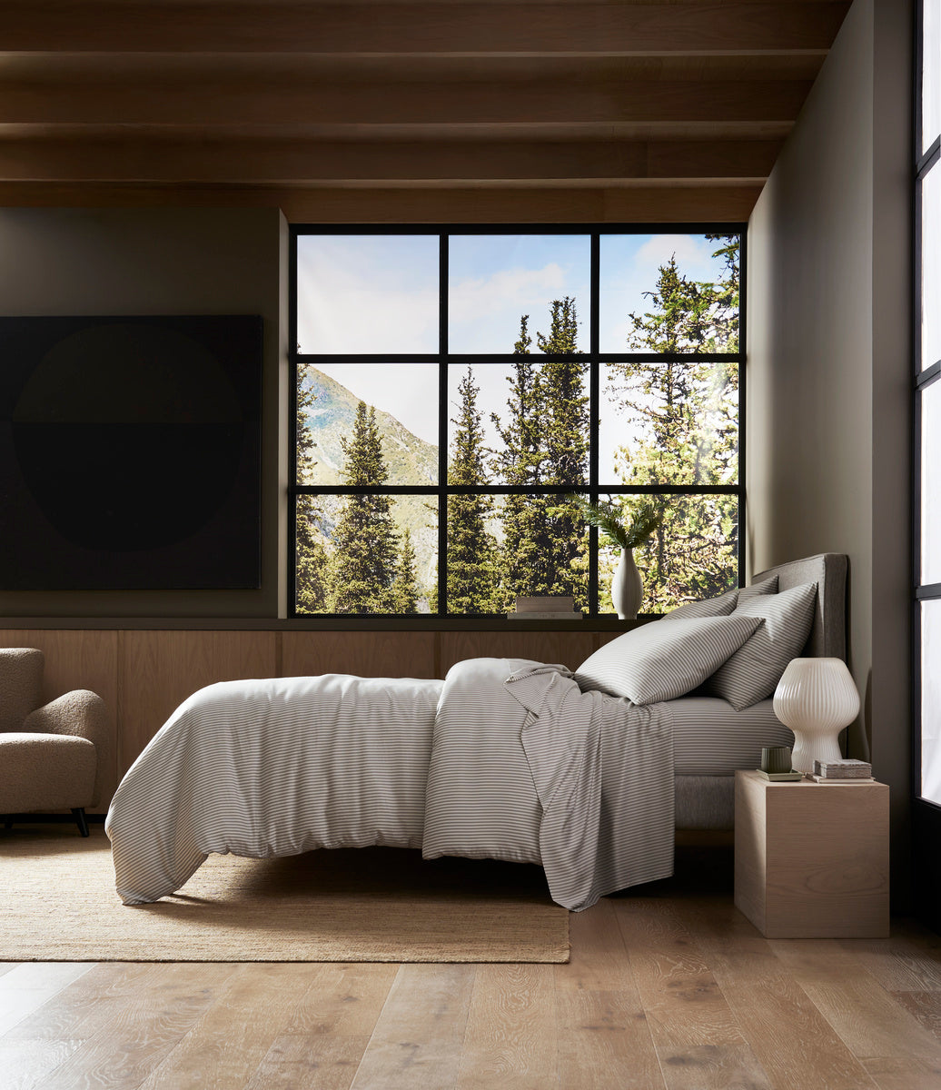 A modern bedroom features a neatly made bed with Cozy Earth's Bamboo Duvet Cover, light bedding, a nightstand and lamp, wood floors, and large windows overlooking pine trees and mountains under a blue sky filled with natural light.|Color:Classic Stripe in Laurel