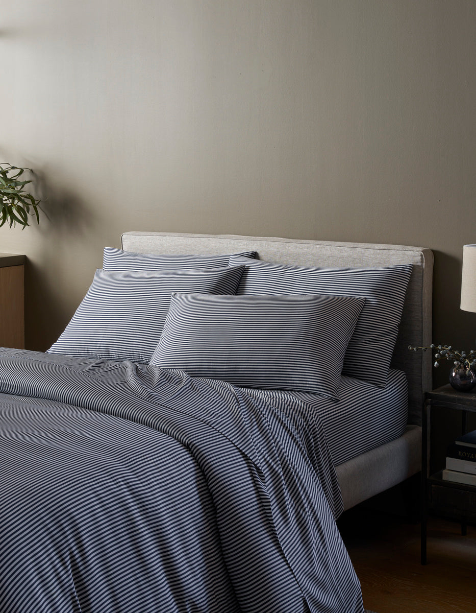 A neatly made bed features the Cozy Earth Bamboo Duvet Cover in gray and white stripes, complemented by matching pillows and a comforter. Against a beige wall, it’s flanked by a nightstand with a lamp, vase, and a plant in the corner.|Color:Classic Stripe in Pacific