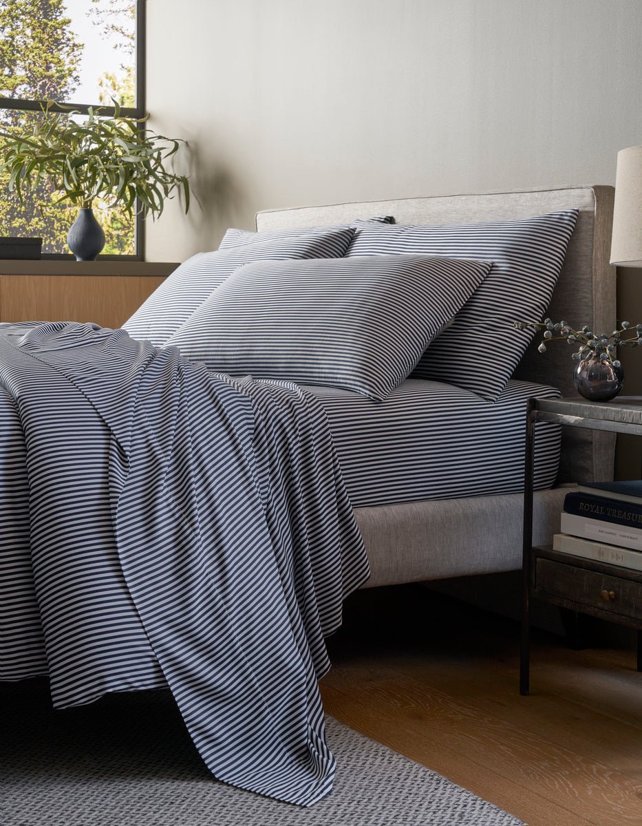 A modern bedroom features a neatly made bed with Cozy Earth’s Bamboo Bedding Core Bundle in black and white stripes, accompanied by a small nightstand holding books, a lamp, and a vase of flowers under soft lighting.|Color:Classic Stripe in Pacific