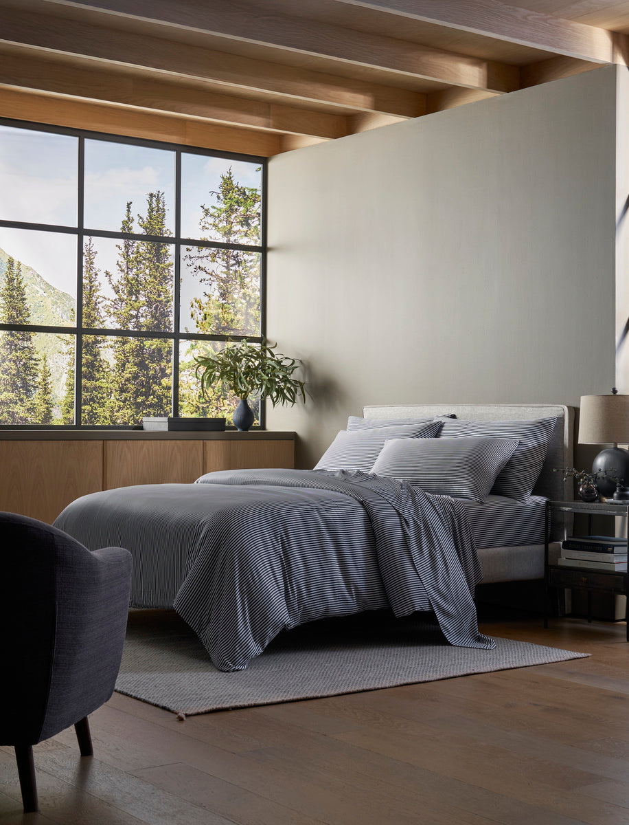 A modern bedroom with a large window, gray armchair, light rug, and wooden floors features a neatly made bed with Cozy Earth's Bamboo Duvet Cover. Natural light fills the space, with plants and decor displayed on the dresser.|Color:Classic Stripe in Pacific