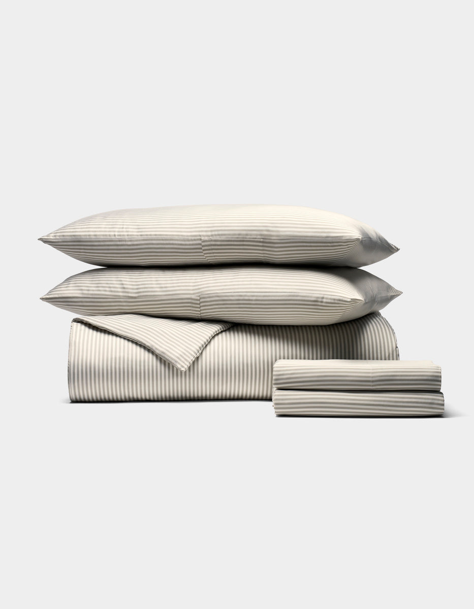 The Bamboo Bedding Deluxe Bundle by Cozy Earth is shown neatly stacked with two pillows, a folded comforter, and sheets in a subtle light gray and white pinstripe pattern against a plain background.|Color:Classic Stripe in Laurel