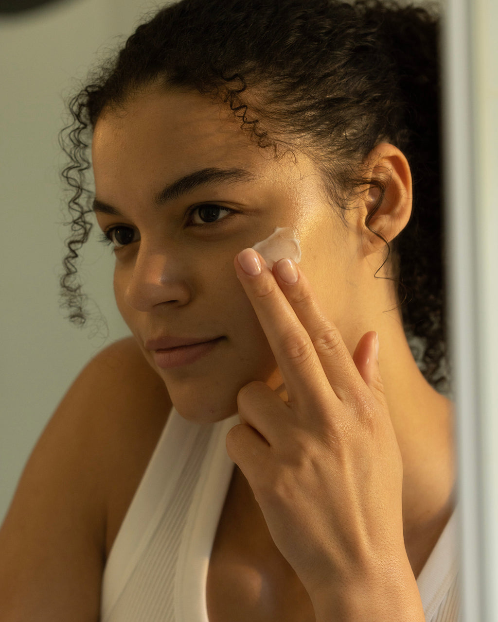 Smiling slightly, a person with curly hair applies Cozy Earth's Water Cream Moisturizer to their face with two fingers. They're wearing a white tank top, and the soft lighting creates a calm ambiance as they look to the side. |Size:Mini