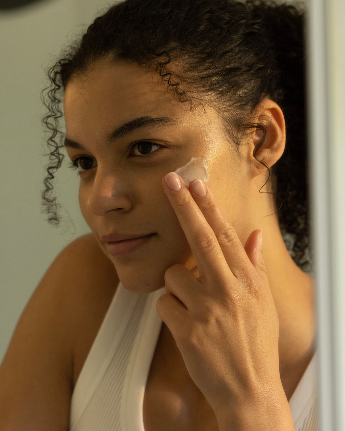 Smiling slightly, a person with curly hair applies Cozy Earth's Water Cream Moisturizer to their face with two fingers. They're wearing a white tank top, and the soft lighting creates a calm ambiance as they look to the side. |Size:Mini