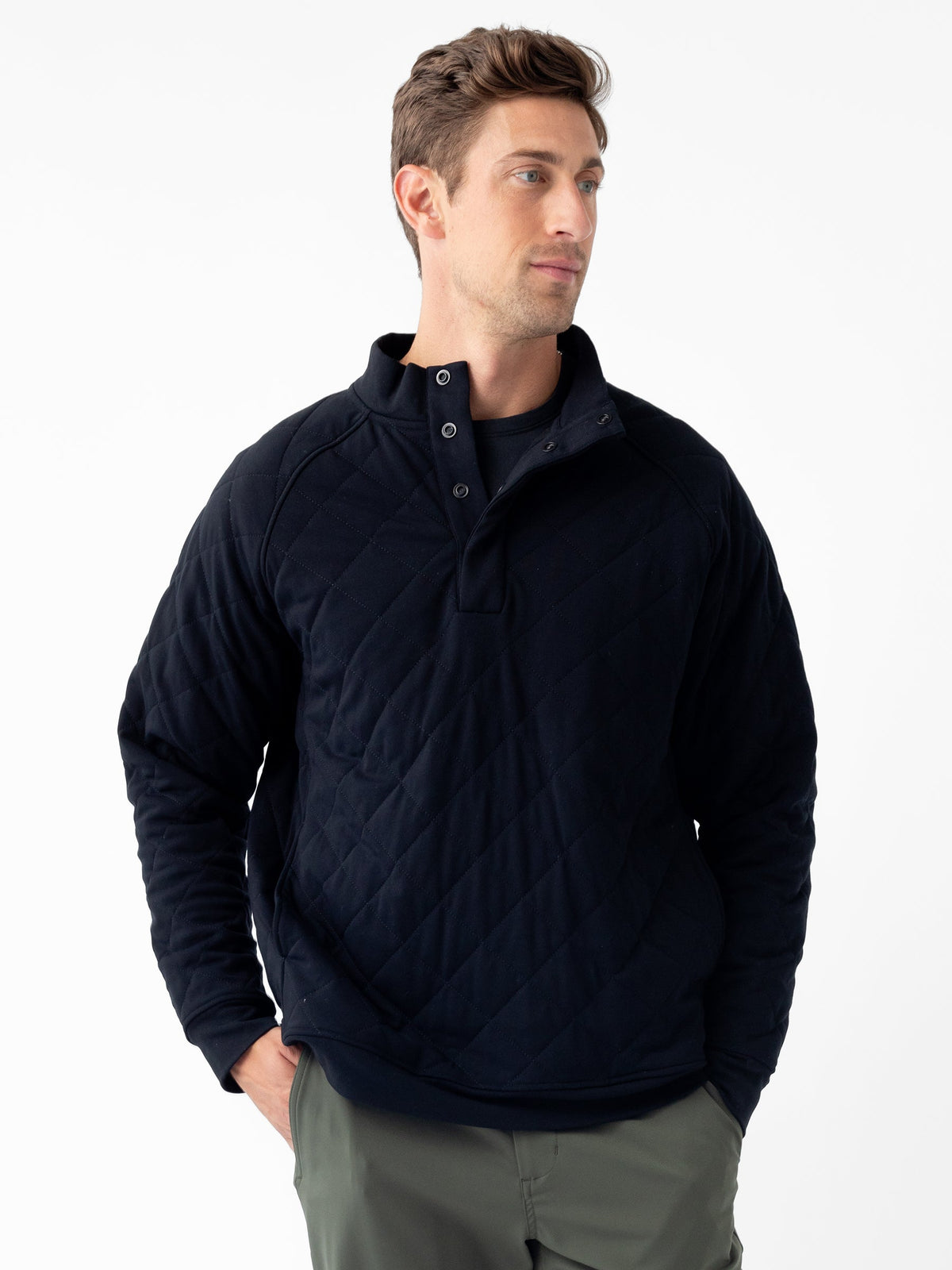 A person stands against a white background wearing Cozy Earth's Men's Ultra-Soft Bamboo Quilted Snap Pullover, which features a high collar and button placket. Their hands are tucked into the pockets of their gray pants, and they are looking slightly to the left with a neutral expression. |Color:Black
