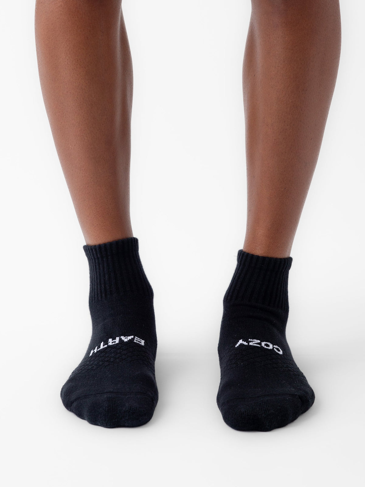 A person wears black Cozy Earth Essential Quarter Socks on a white surface. The socks have white text: "EARTH" on the left toe and "COZY" on the right. |Color:Black