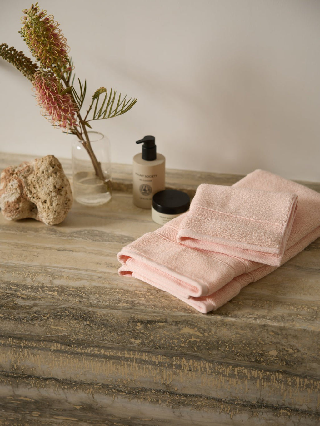 A neatly folded Luxe Washcloth set by Cozy Earth rests on a marble countertop. Beside it are a natural sponge, a small potted plant with red spiky flowers, and two bottles of skincare products, creating a spa-like atmosphere. |Color:Blossom