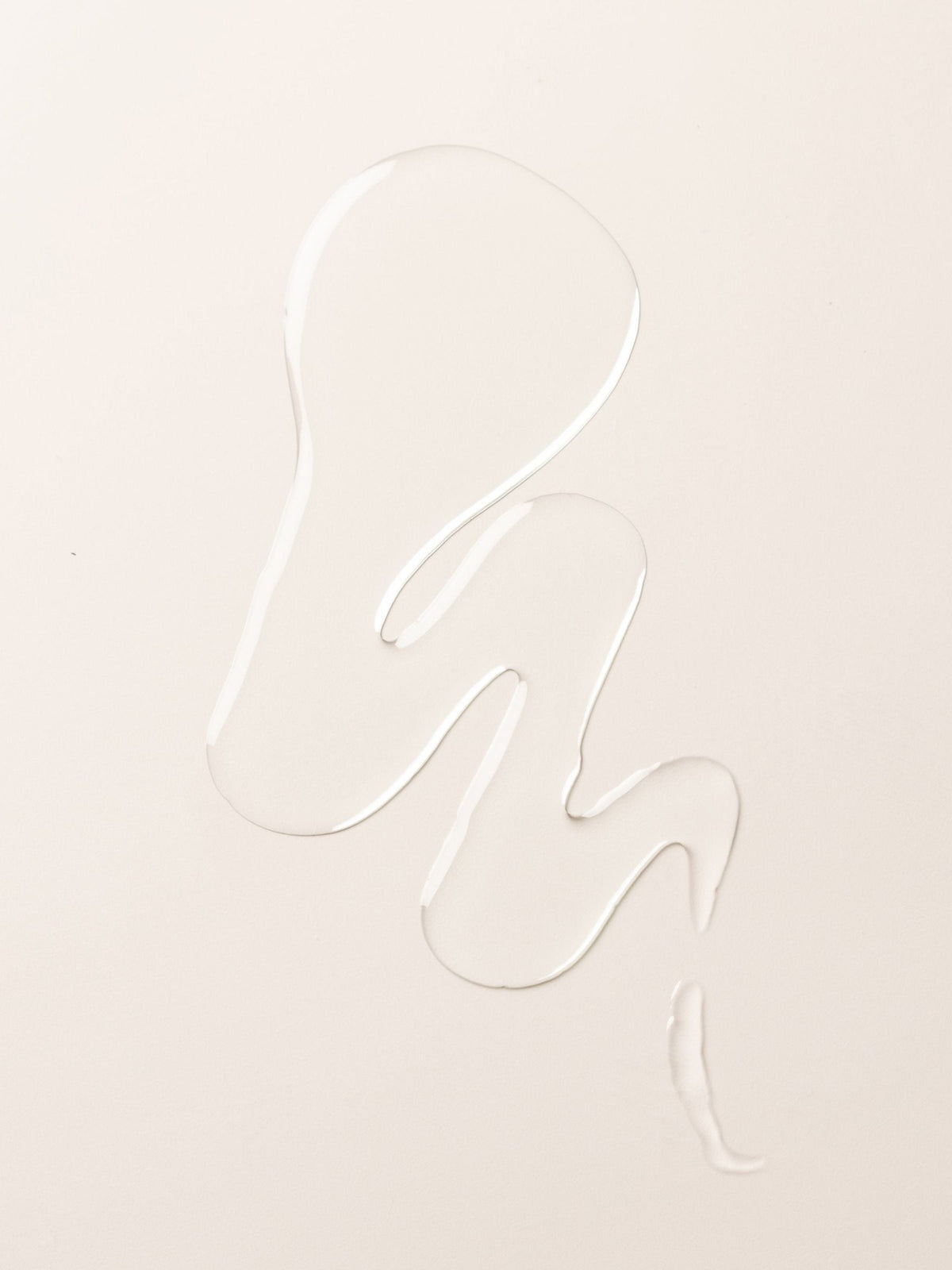 A clear liquid body oil from Cozy Earth forms an abstract, wavy shape on a smooth, off-white surface.
