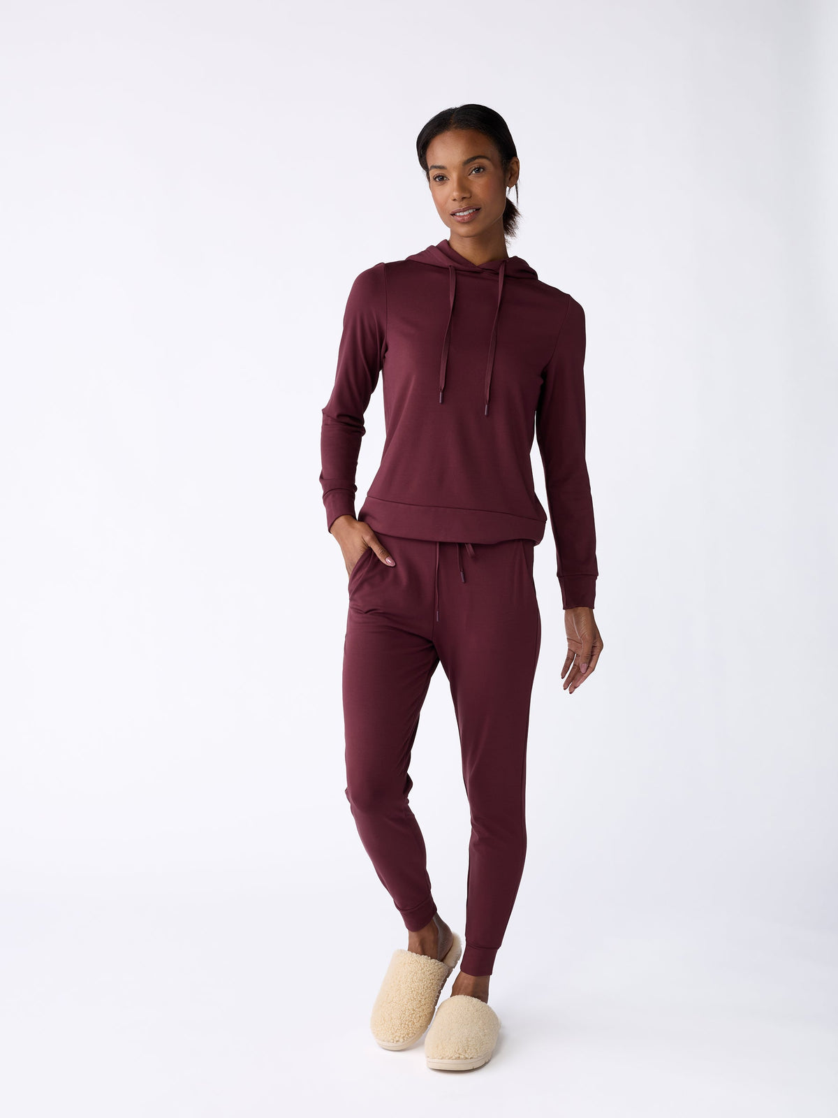 A person wearing Cozy Earth's maroon Women’s Bamboo Jogger Pant and a matching hoodie stands against a plain white background. They have their hands in their pockets and are wearing beige slippers. |Color:Burgundy