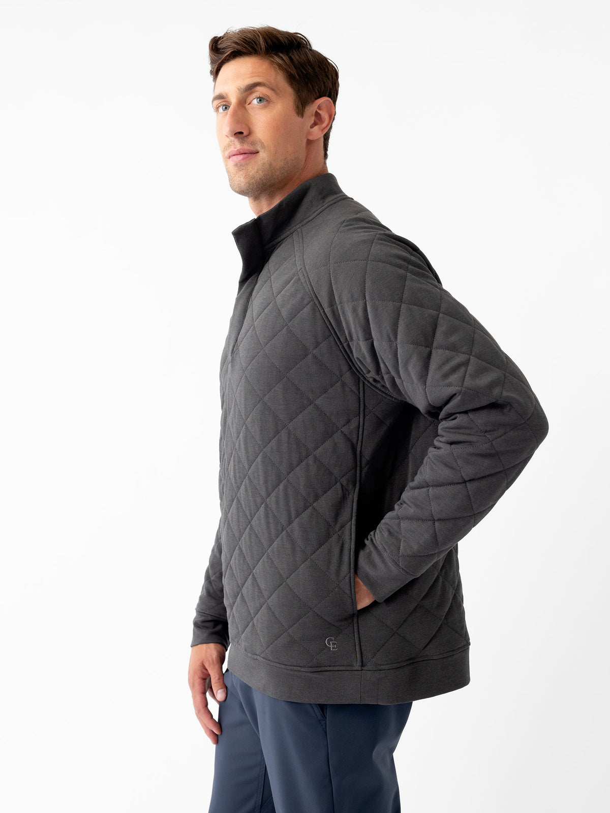 A man wearing the Men's Ultra-Soft Bamboo Quilted Snap Pullover in dark gray by Cozy Earth and dark blue pants stands against a plain white background. He is posing with both hands in his pockets and looking slightly to his left. The pullover features a high collar and a quilted pattern. |Color:Charcoal