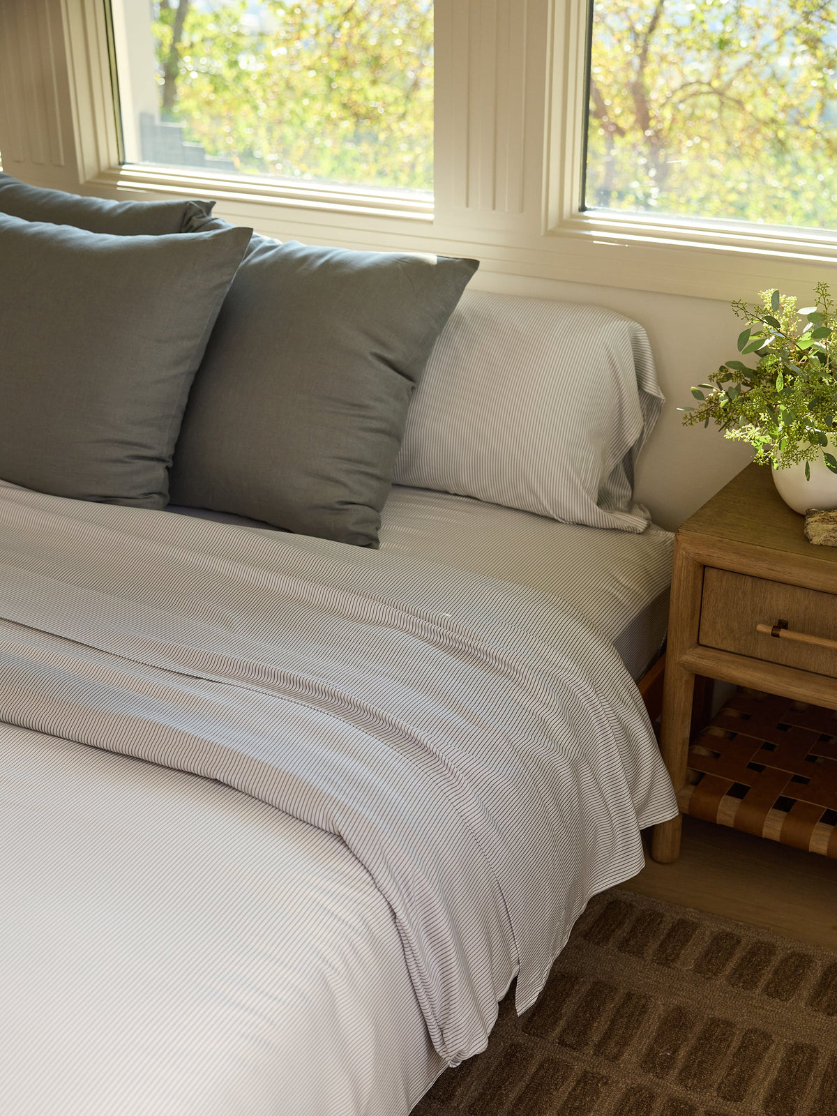 A neatly made bed showcases the Cozy Earth Bamboo Duvet Cover, designed with white and gray stripes, complemented by three gray pillows. A wooden nightstand holds a small plant, while sunlight streams through the windows, casting a warm glow on the bed. A brown woven rug is partially visible on the floor. |Color:Charcoal Pinstripe