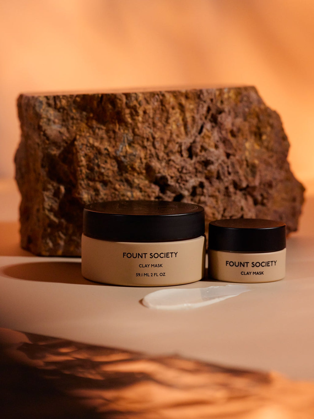 Two jars of Cozy Earth's Clay Mask sit on a beige surface with a textural brown rock backdrop. A clay mask smear is visible in front, highlighting a natural, earthy theme. |Size:Mini