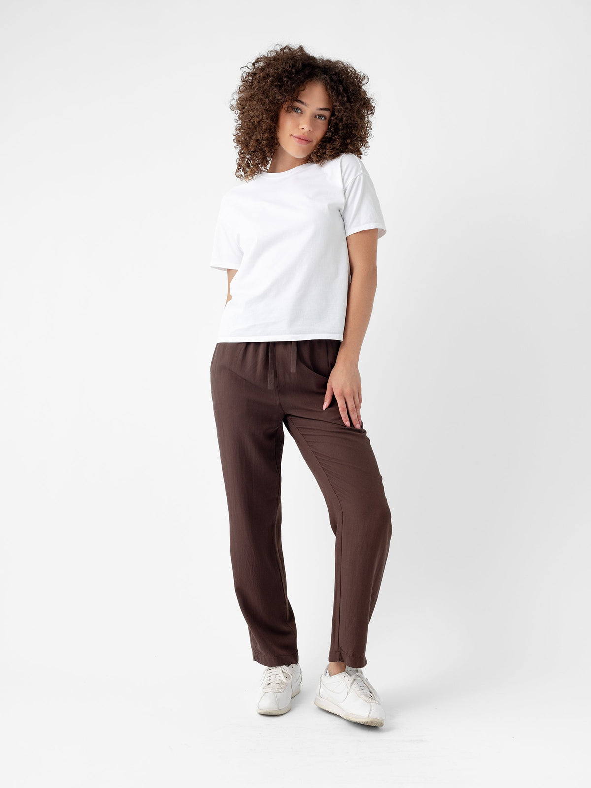 A person with curly hair stands against a plain white background, wearing a white T-shirt, Women's Sunset Cropped Pant from Cozy Earth, and white sneakers. They have a relaxed posture with one hand in their pocket and a calm expression. |Color:Cacao
