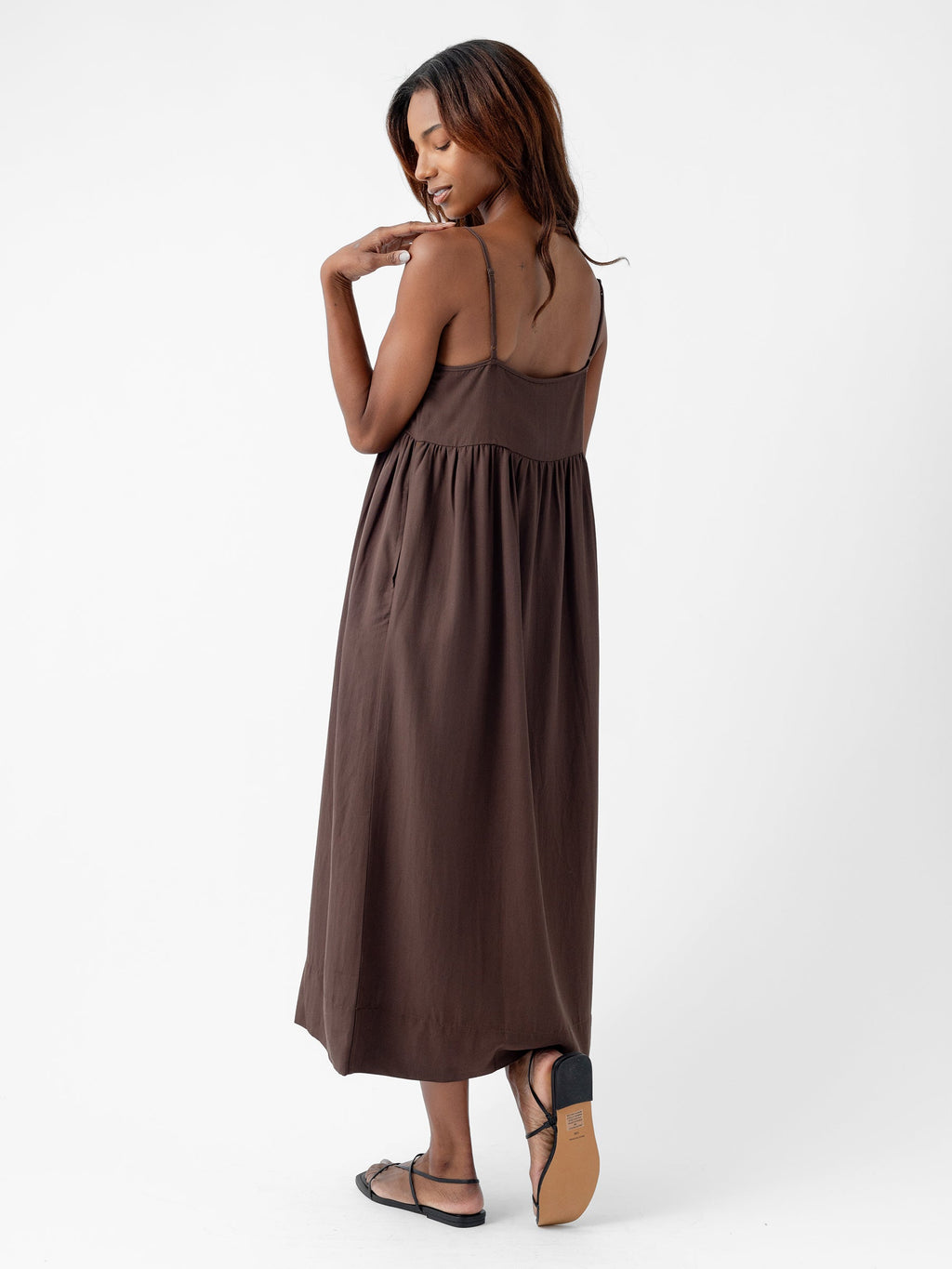 A woman with long hair touches her shoulder as she looks down, wearing Cozy Earth's Women's Sunset Gathered Dress, a sleeveless, brown maxi dress with spaghetti straps and black sandals. She is standing on a white background, posing with one foot slightly raised. |Color:Cacao