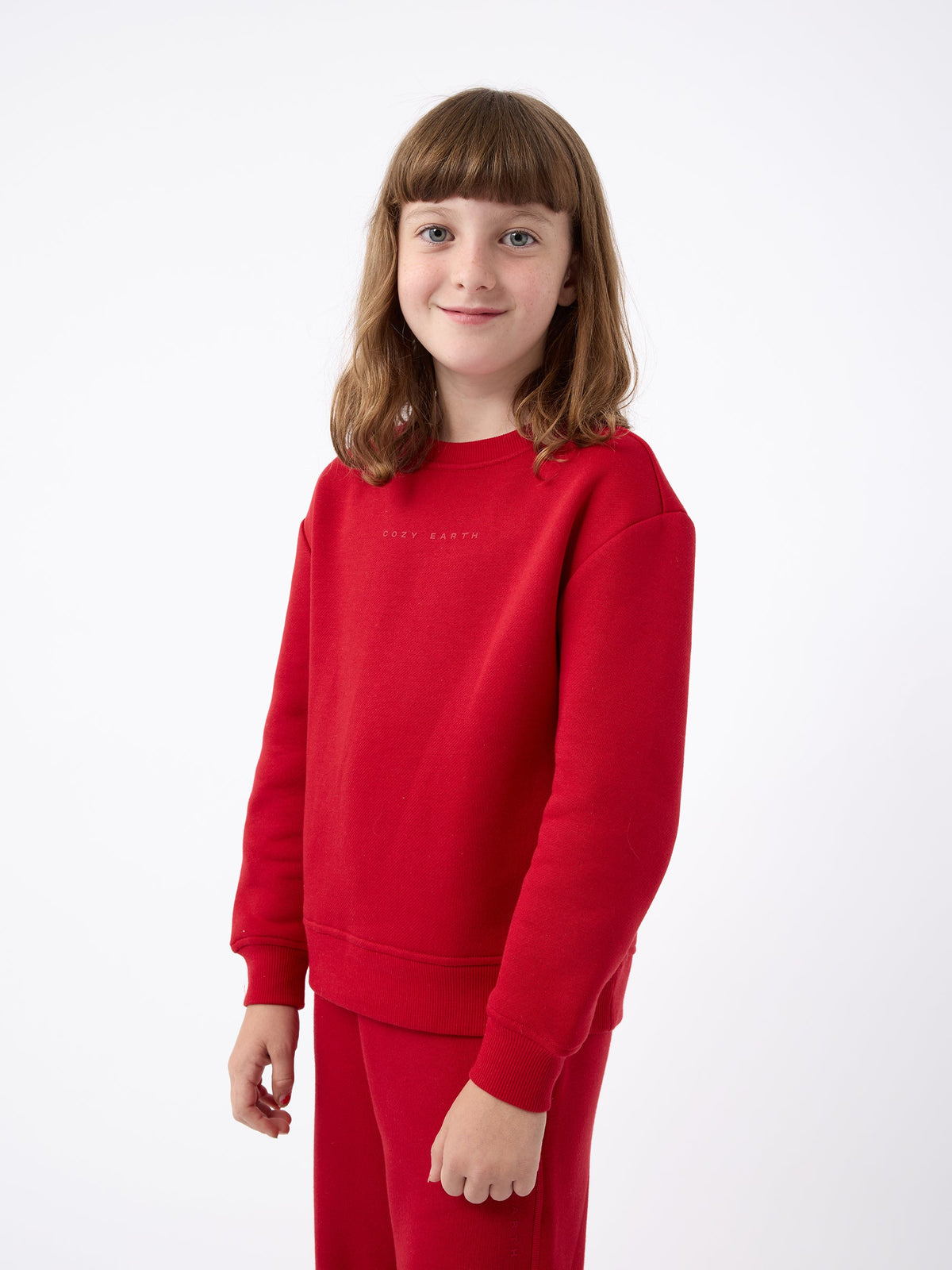 A cheerful young child with brown hair stands against a simple white background, sporting the Kid's CityScape Crewneck and matching pants from Cozy Earth. |Color:Crimson