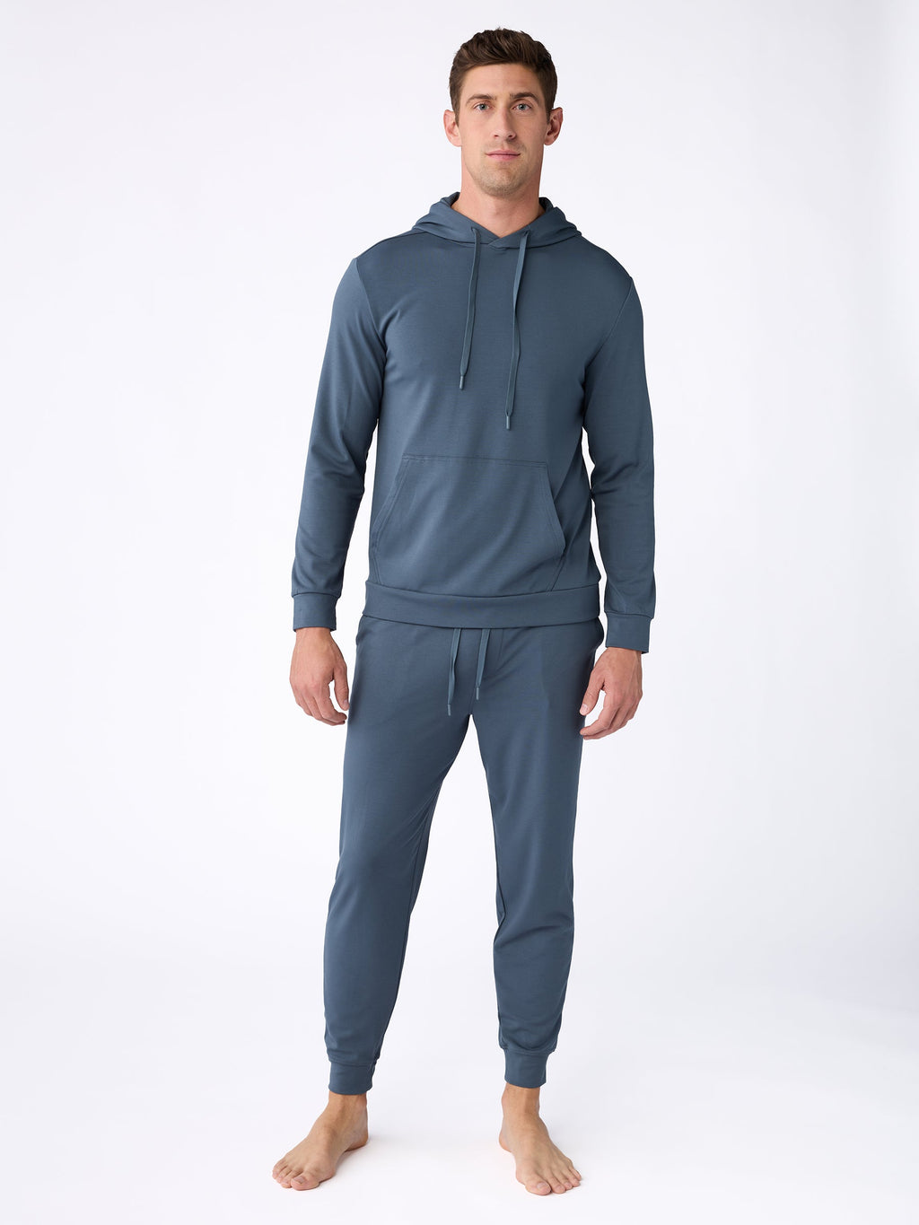 A man with short, dark hair and a neutral expression stands barefoot on a white background, dressed in Cozy Earth's Men's Ultra-Soft Bamboo Pullover Crew in teal. The pullover features a front pocket and drawstrings, while the matching jogger pants are fitted and comfortable. |Color:Dawn
