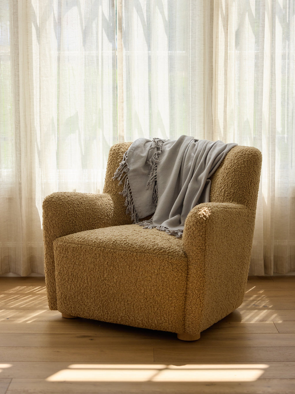 A cozy beige armchair with textured fabric is placed in front of a sheer, sunlit curtain. A light gray Bamboo Tassel Throw by Cozy Earth is draped casually over the backrest, and sunlight casts soft shadows on the wooden floor. |Color:Dove Grey