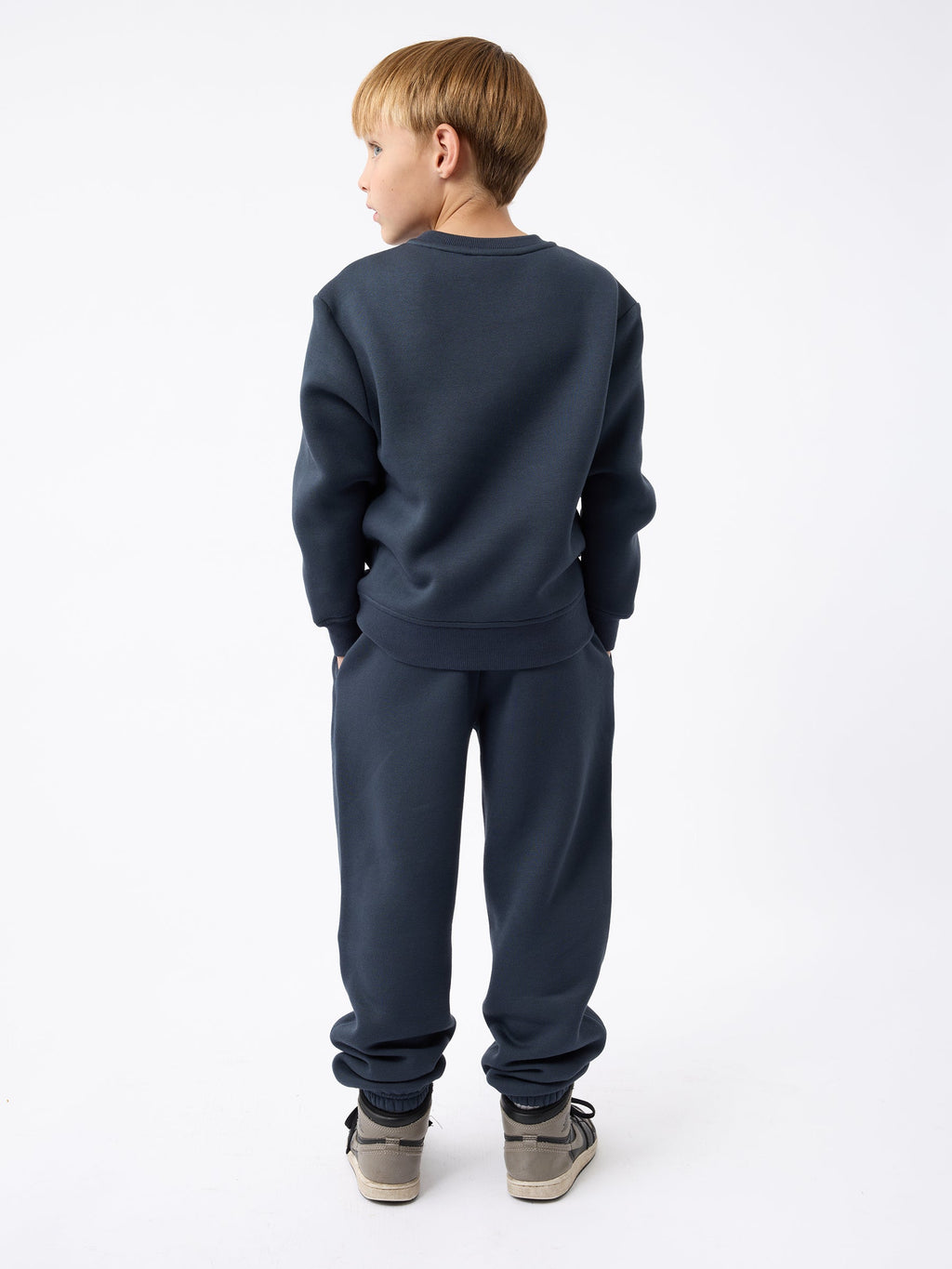 A young boy with blonde hair is standing with his back to the camera, wearing a Kid's CityScape Crewneck in dark blue from Cozy Earth and gray sneakers. His hands are in his pockets, and he is looking slightly to the side against a plain white background. |Color:Eclipse