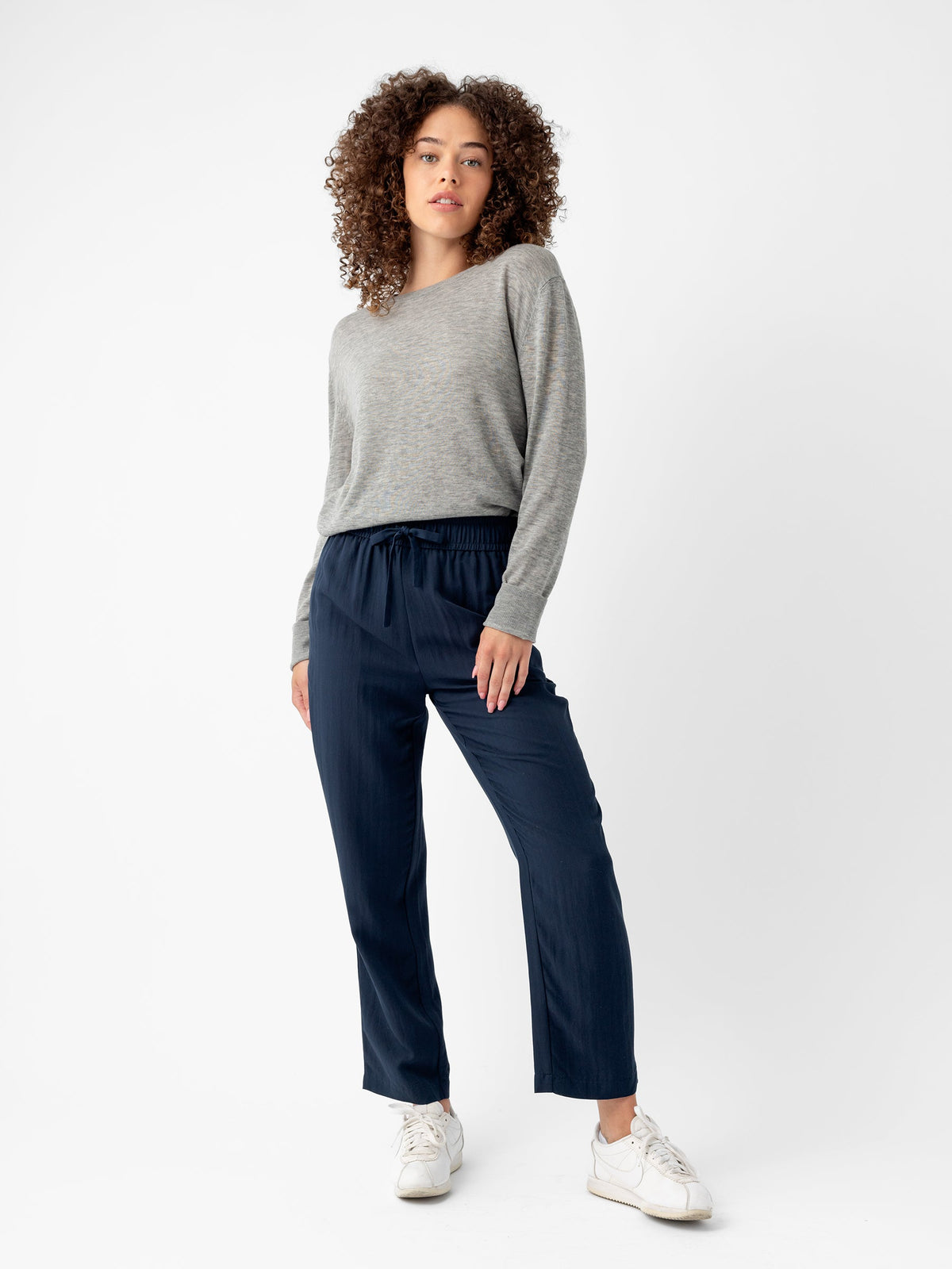A woman with curly hair is standing against a plain white background. She is wearing a light grey long-sleeve shirt, Cozy Earth’s dark blue Women's Sunset Cropped Pant, and white sneakers. She has a relaxed pose with one hand in her pocket and a neutral expression on her face. |Color:Eclipse