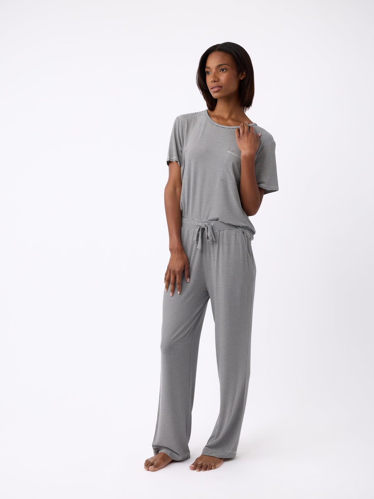 A woman stands barefoot against a white background, her hand raised to her neck. She's wearing Cozy Earth's loose-fitting Women's Bamboo Stretch-Knit Pant in gray, paired with a matching T-shirt. |Color:Eclipse Mini Stripe