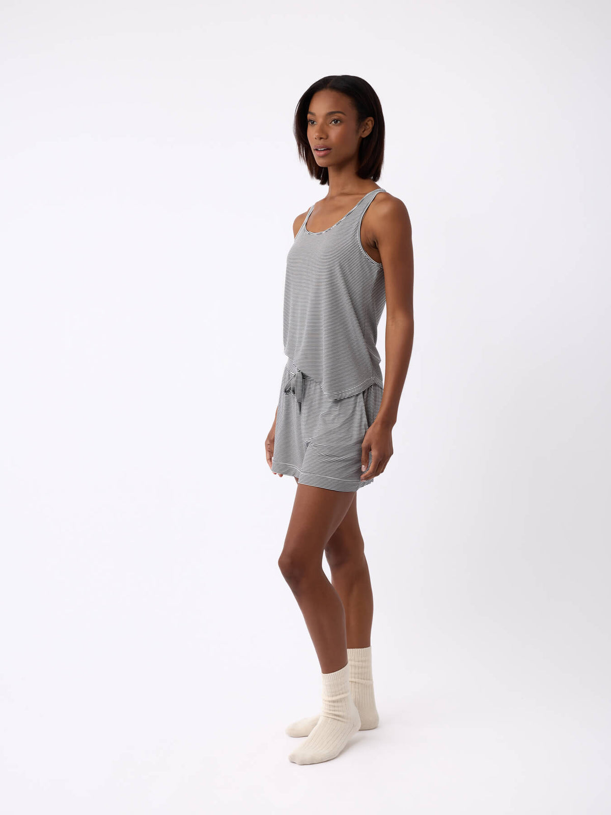 A person with shoulder-length hair stands against a plain background, wearing Cozy Earth's Women's Bamboo Stretch-Knit Lounge Tank in gray and matching shorts. They are also wearing beige socks and facing slightly to the side. |Color:Eclipse Mini Stripe