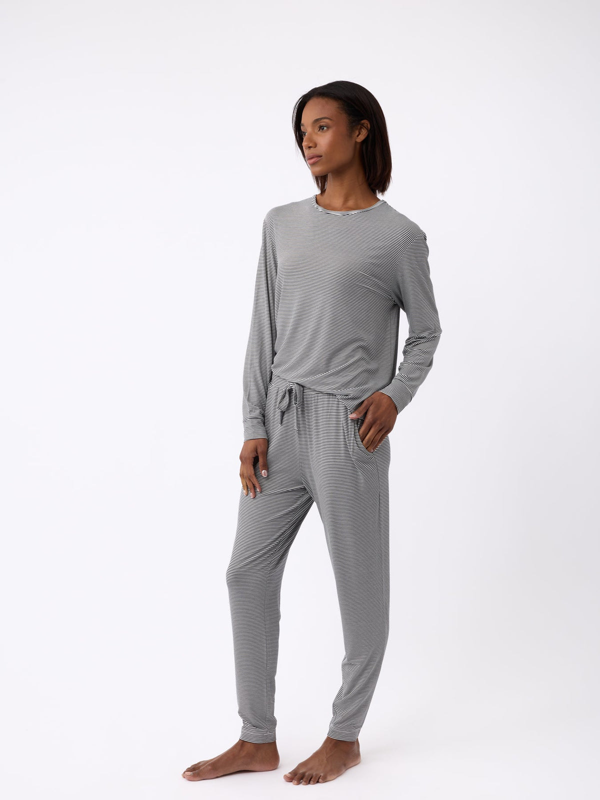 A person in Cozy Earth's Women's Bamboo Stretch-Knit Tapered Pant, gray and soft-looking with pockets, stands barefoot against a white background. They have one hand in a pocket and are gazing to the side. |Color:Eclipse Mini Stripe