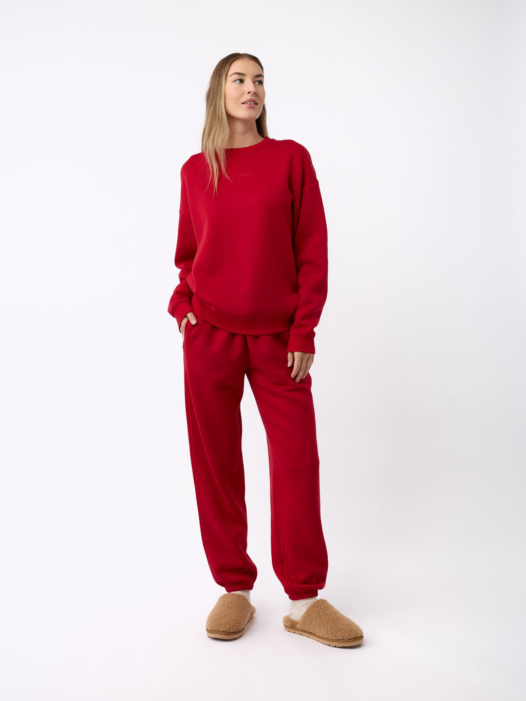 A person stands against a plain background, wearing a matching red sweatshirt and sweatpants set. They have light brown hair and are gazing to the side. On their feet, they are wearing Cozy Earth's Puffy Sheep Slippers in tan, which look fuzzy and comfortable. They have one hand in their pocket. |Color:Fawn