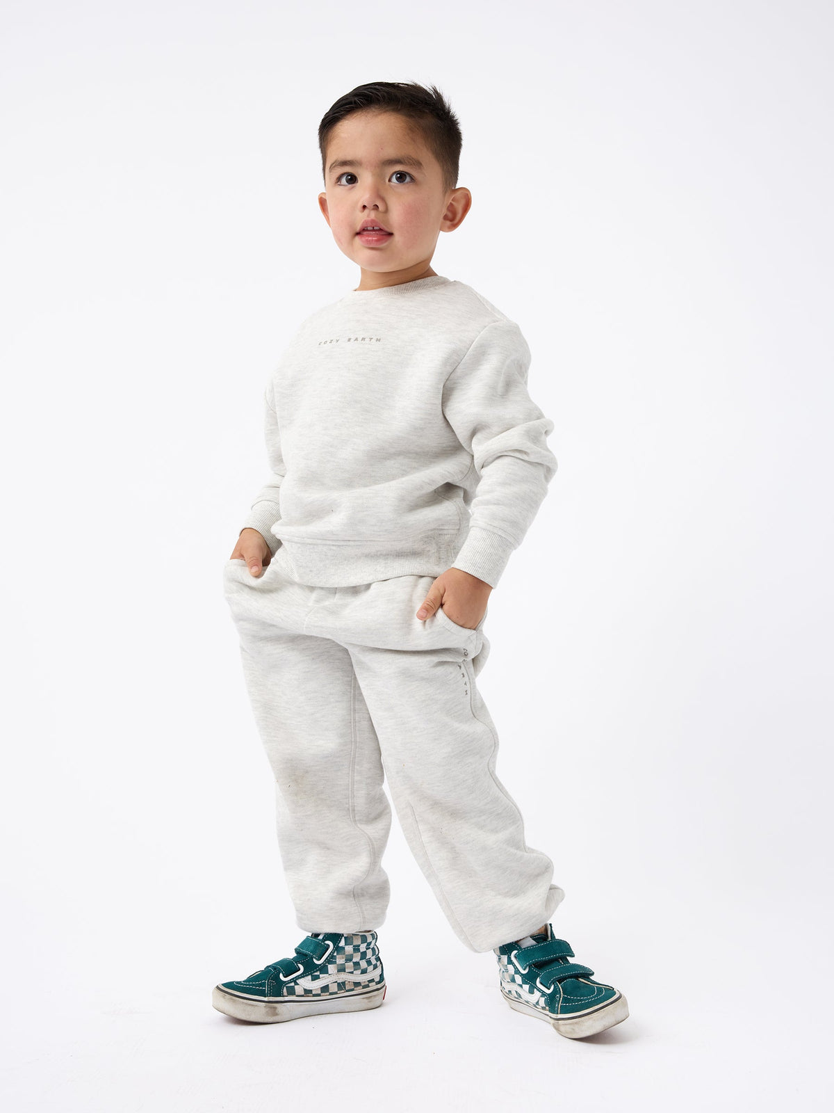 A young child stands confidently against a plain background, dressed in the Kid's CityScape Crewneck by Cozy Earth and sporting green sneakers. With hands tucked into pockets, the child strikes a relaxed pose. The outfit looks comfortable and casual. |Color:Heather Grey