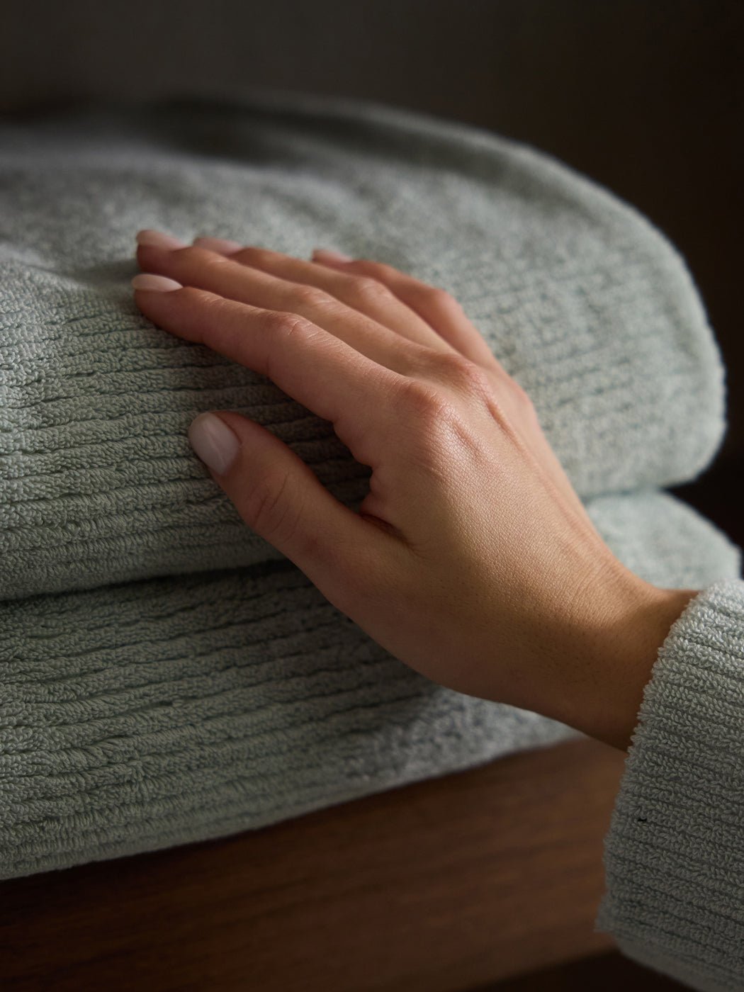 A hand gently rests on a neatly folded Ribbed Terry Bath Sheet by Cozy Earth, showcasing its textured gray fabric on a wooden surface.|Color:Laurel