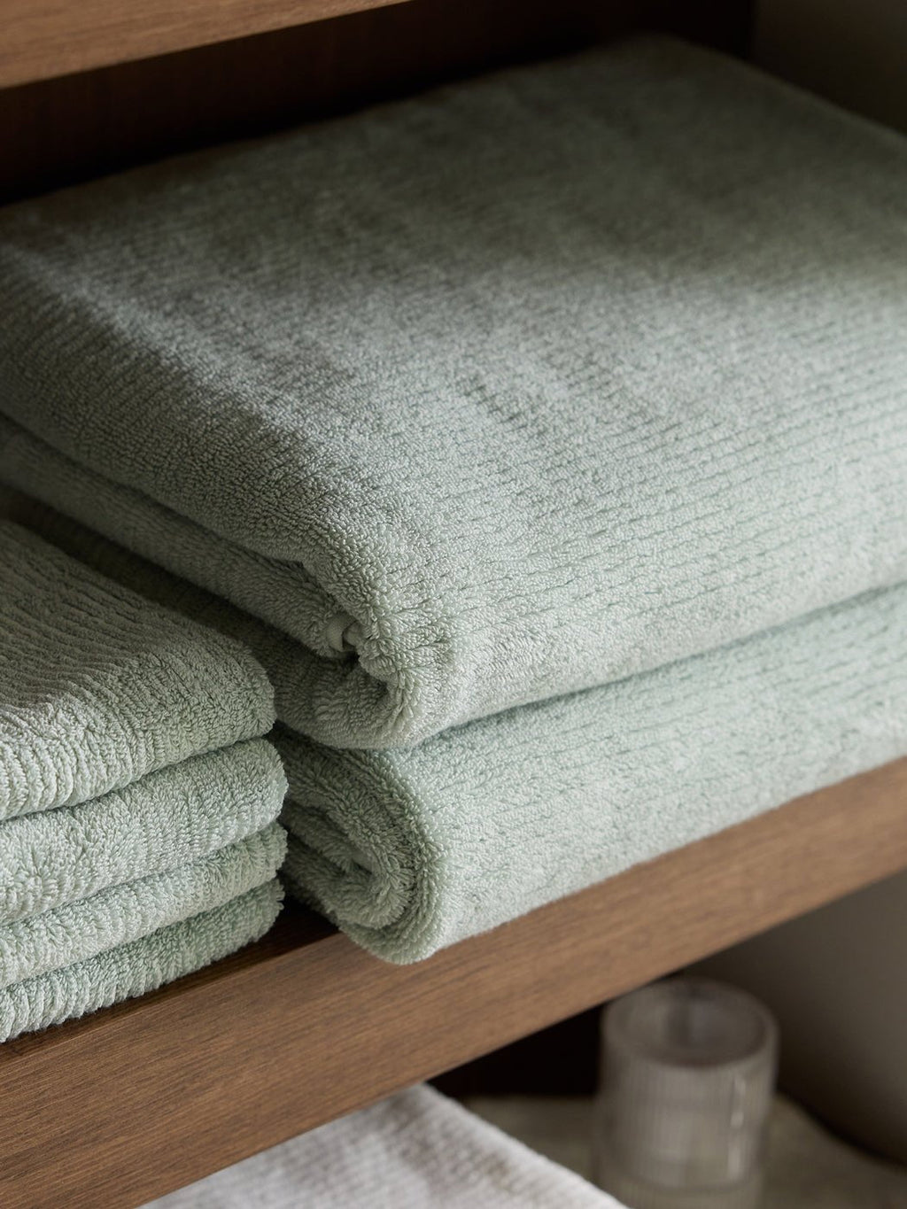 Two neatly folded Ribbed Terry Bath Towels by Cozy Earth in light green rest on a wooden shelf, with more towels and a frosted glass container nearby.|Color:Laurel
