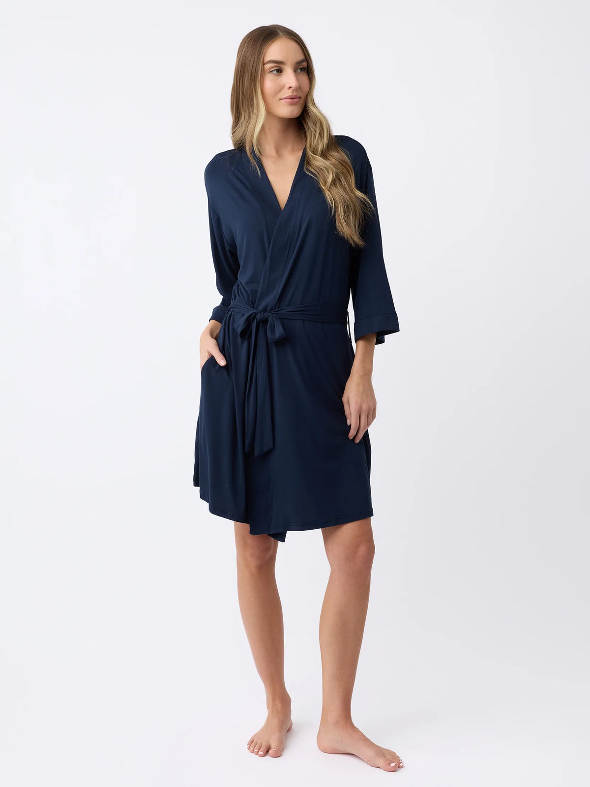 A person with long hair poses barefoot against a plain white background, wearing Cozy Earth's Women's Stretch-Knit Bamboo Kimono Robe in navy blue, featuring long sleeves, a wrap style, and a waist tie. |Color:Navy