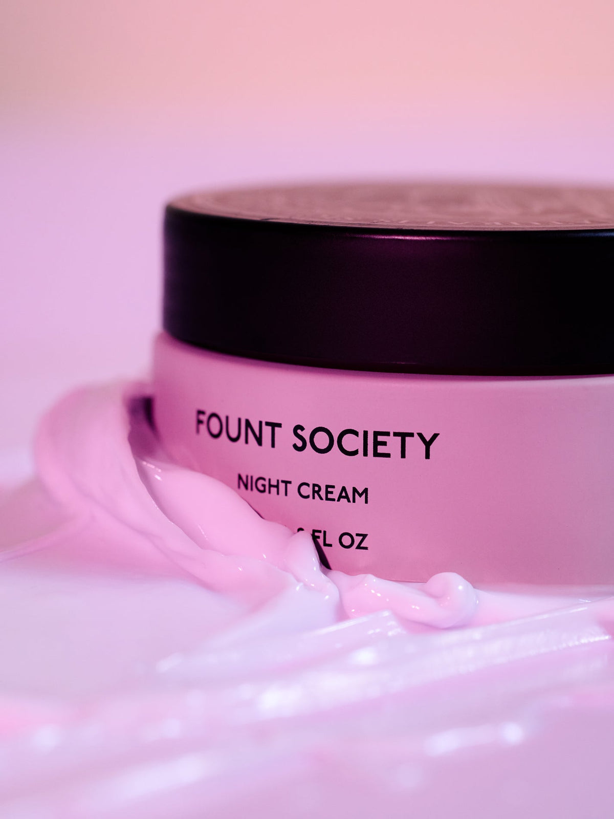 A pink jar of Cozy Earth Night Cream with a black lid, surrounded by swirls of light pink cream, sits against a softly blurred pink background. |Size:Full Size