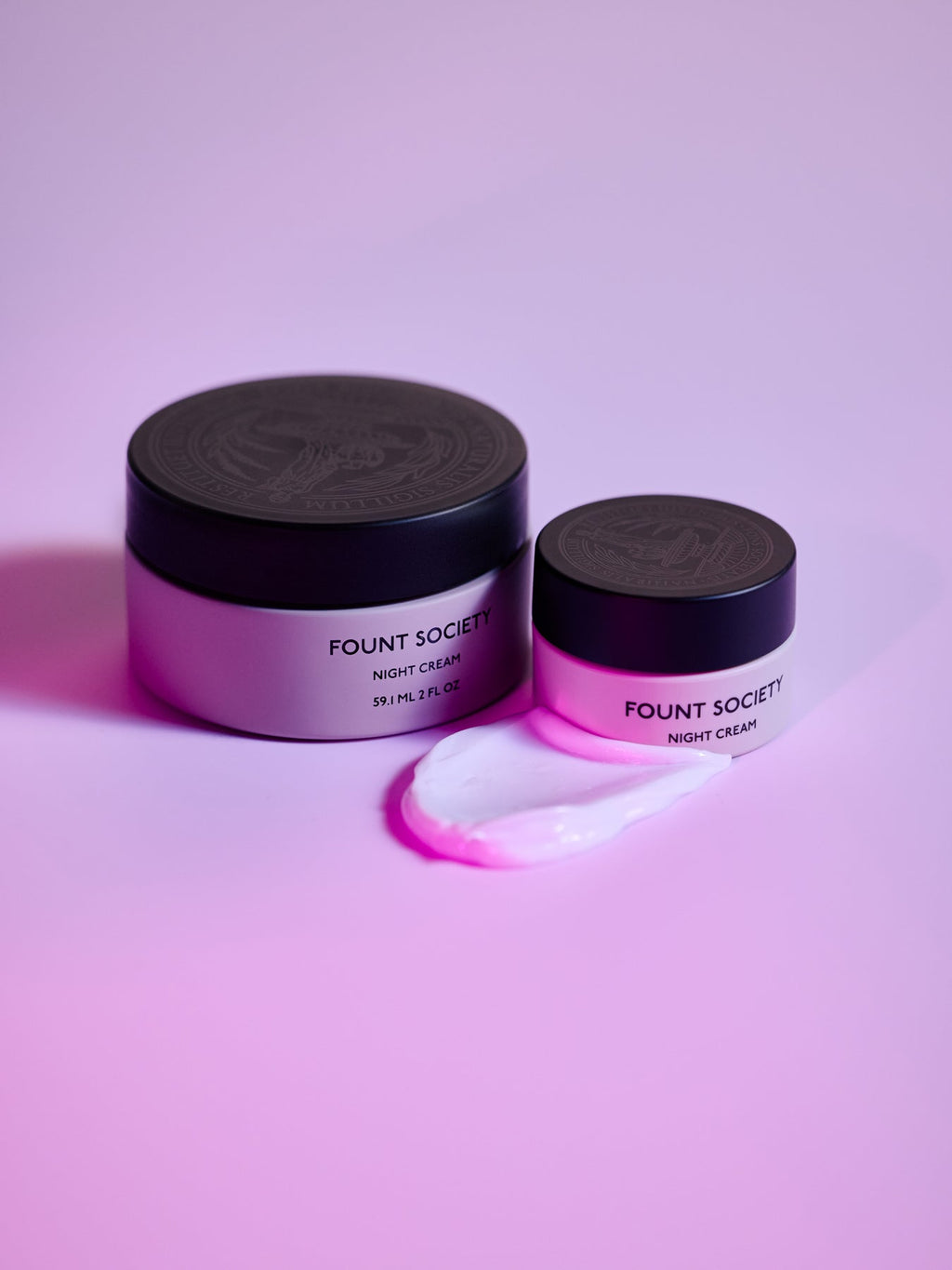 Two jars of Cozy Earth Night Cream rest on a pink surface. The larger jar is partially open, revealing a creamy smear beside the smaller, closed jar. Both have black lids. |Size:Mini