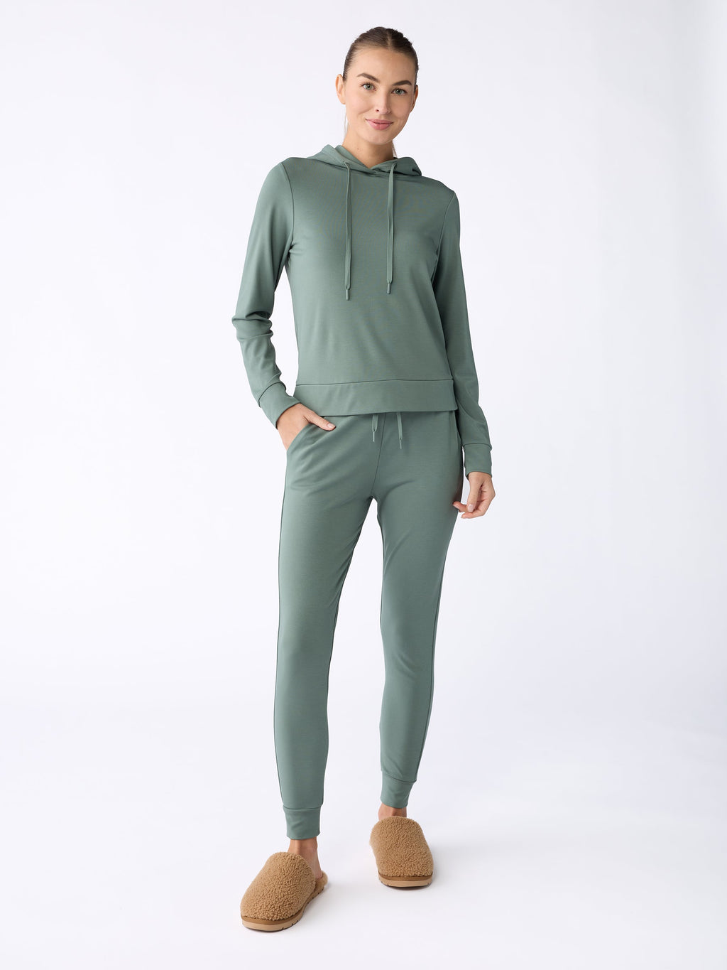 A person dressed in a light green hoodie paired with Cozy Earth's Women's Bamboo Jogger Pant stands against a plain background. They are also wearing brown fuzzy slippers, have their hair tied back, and are smiling slightly. |Color:Oakmoss