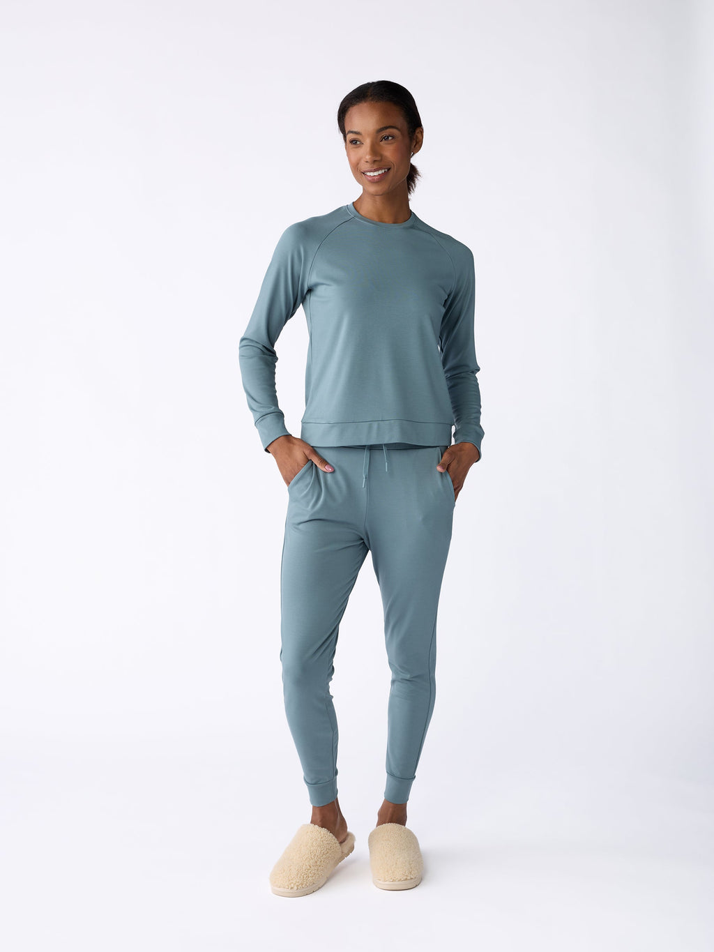 A person in a matching light blue ensemble, including the Women's Bamboo Jogger Pant from Cozy Earth, stands against a plain white background. They accessorize with cream-colored slippers and appear relaxed, with their hands in their pockets. |Color:Oceanside