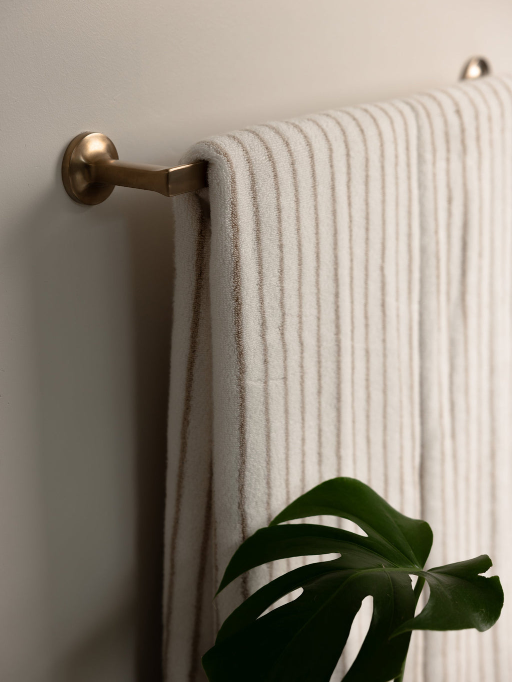 A neatly folded Luxe Bath Towel in textured beige from Cozy Earth hangs on a brass towel bar mounted on a light-colored wall. Below the towel, a green Monstera plant leaf is partially visible, adding a touch of nature to the clean and minimal bathroom setting. |Color:Sand Stripe