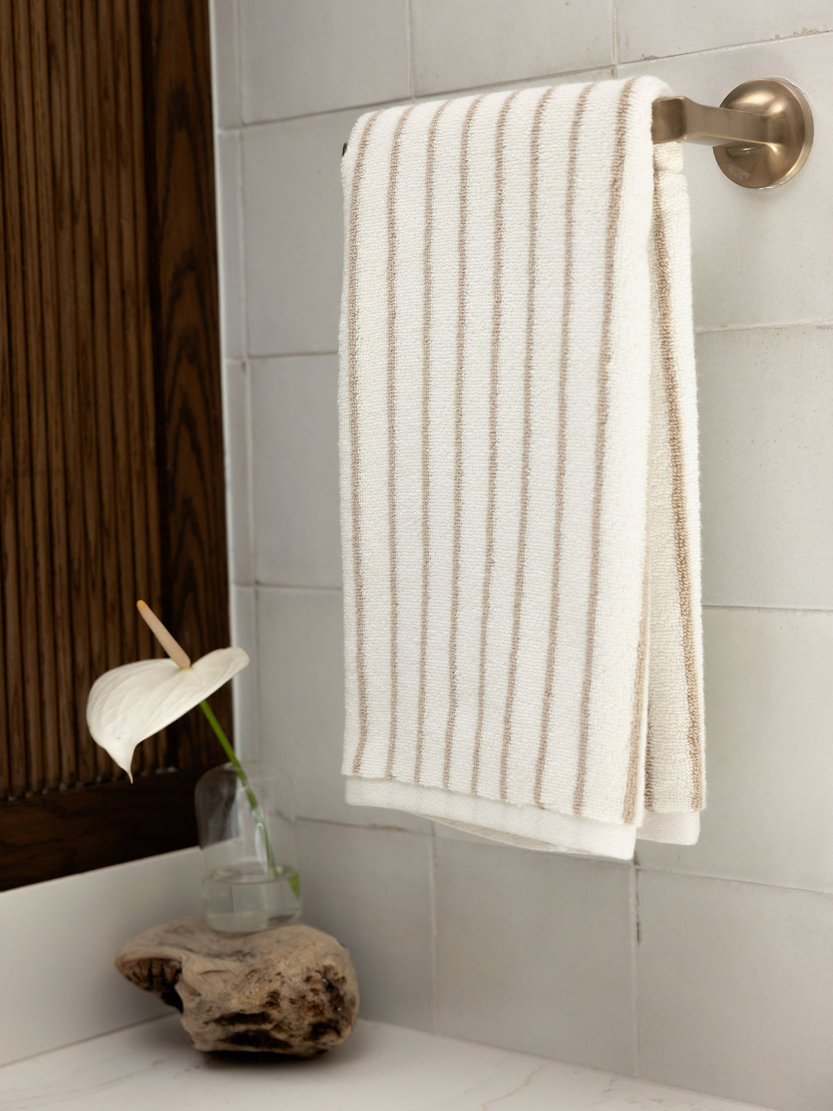 A neatly folded Luxe Hand Towel by Cozy Earth, featuring thin beige stripes, hangs on a silver towel bar in a bathroom. Below, a rustic wooden shelf holds a small glass vase with a single white anthurium flower. The bathroom walls are tiled with large, light-colored tiles. |Color:Sand Stripe