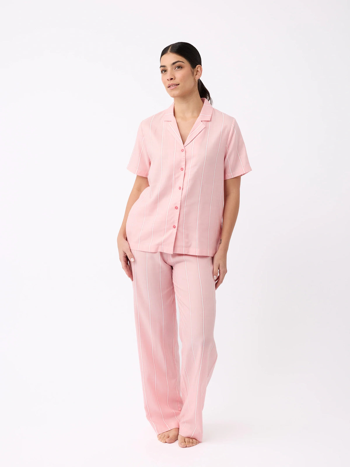 A person models a light pink, pinstriped pajama set from Cozy Earth. The short-sleeve top features buttons and the pants are relaxed fit, all posed against a white backdrop. |Color:Sweetheart Stripe